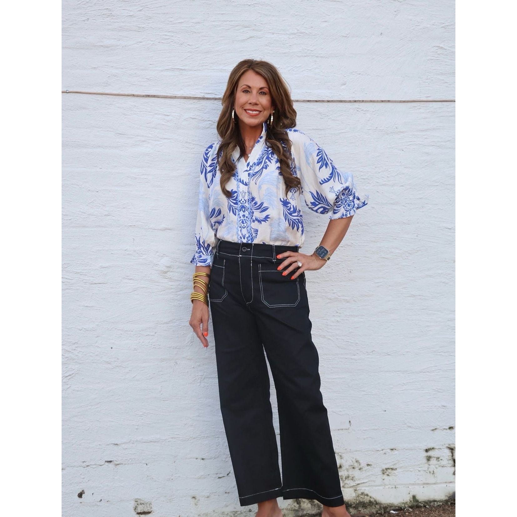 Woman wearing a blue floral blouse and black pants against a white brick wall.