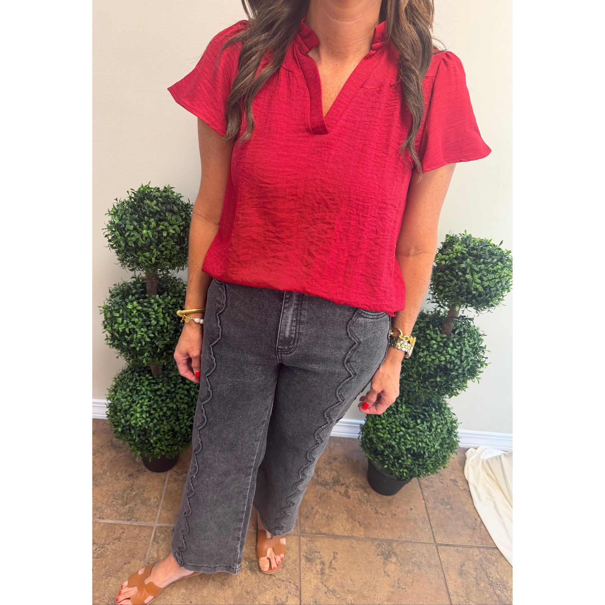 A woman wearing a bright red short-sleeve crinkled blouse and dark gray wide-leg jeans with decorative stitching, standing indoors between two potted green topiary trees.