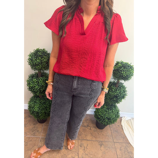 A woman wearing a bright red short-sleeve crinkled blouse and dark gray wide-leg jeans with decorative stitching, standing indoors between two potted green topiary trees.