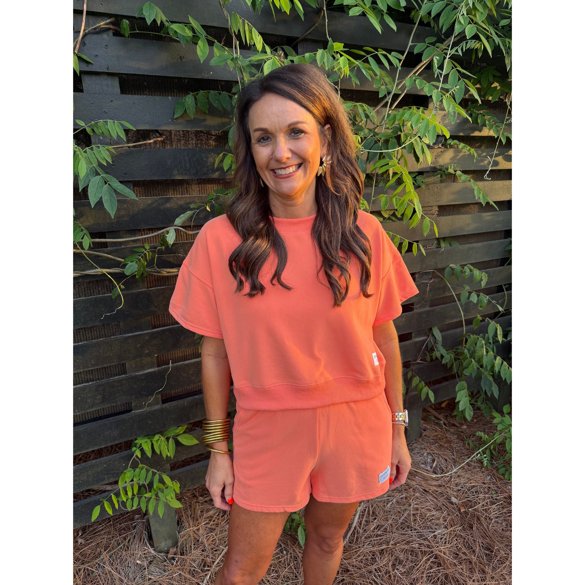 Woman wearing a pink sweatshirt with a logo, standing outdoors with plants in the background