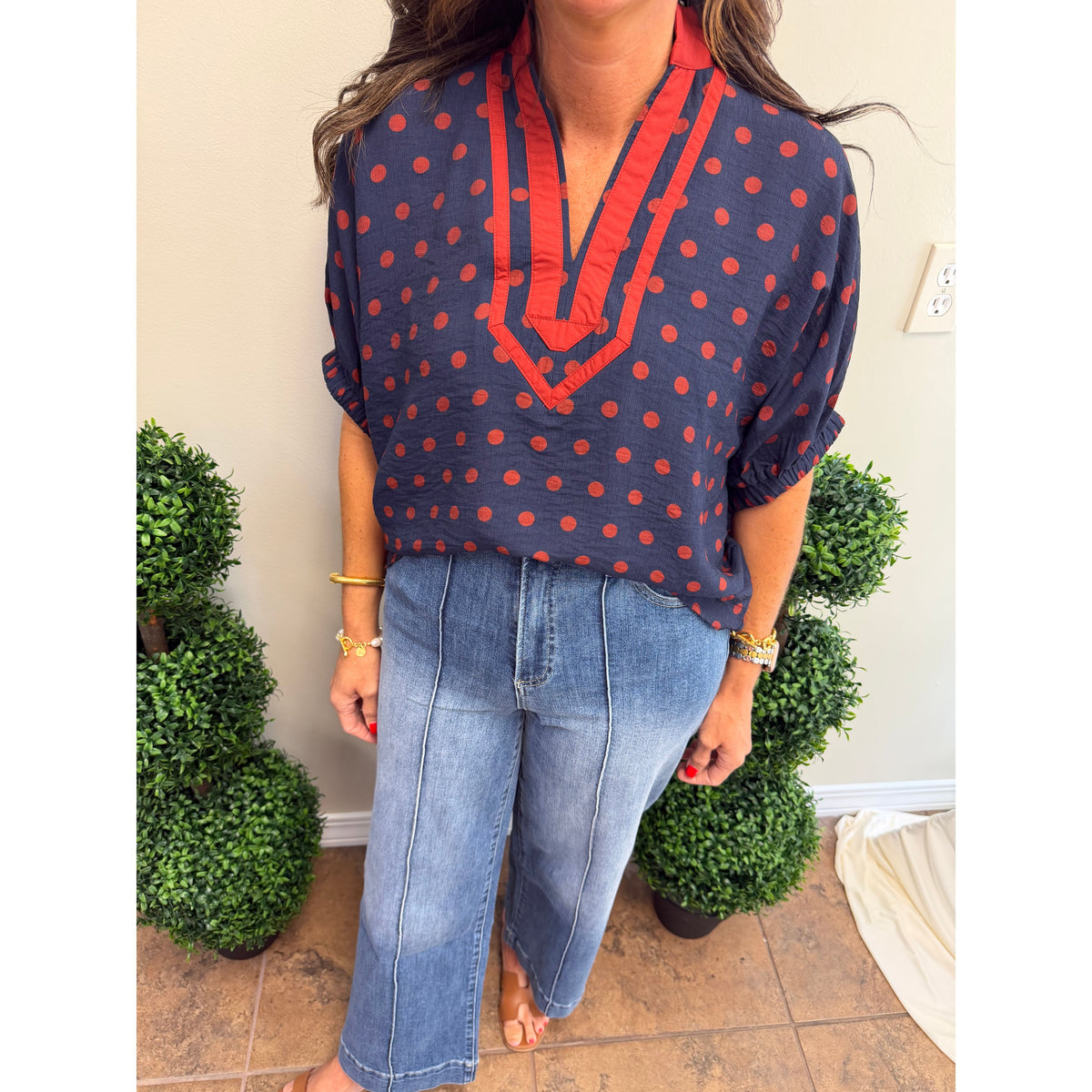 A woman wearing a navy blue blouse with red polka dots and a red ruffled V-neckline, paired with light-wash blue jeans, standing indoors between green potted plants.