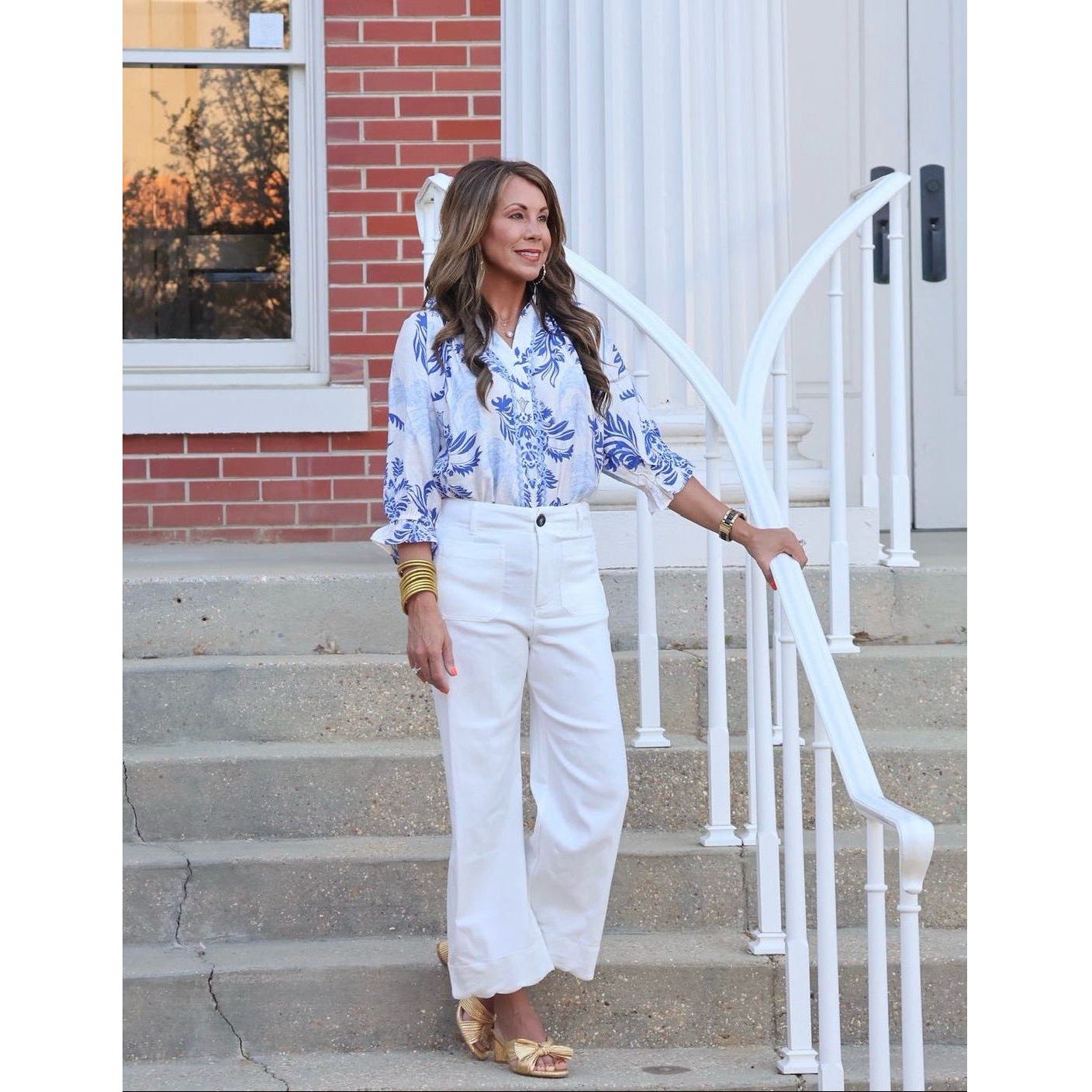 Woman in a blue floral blouse and white pants standing on steps outside a building.