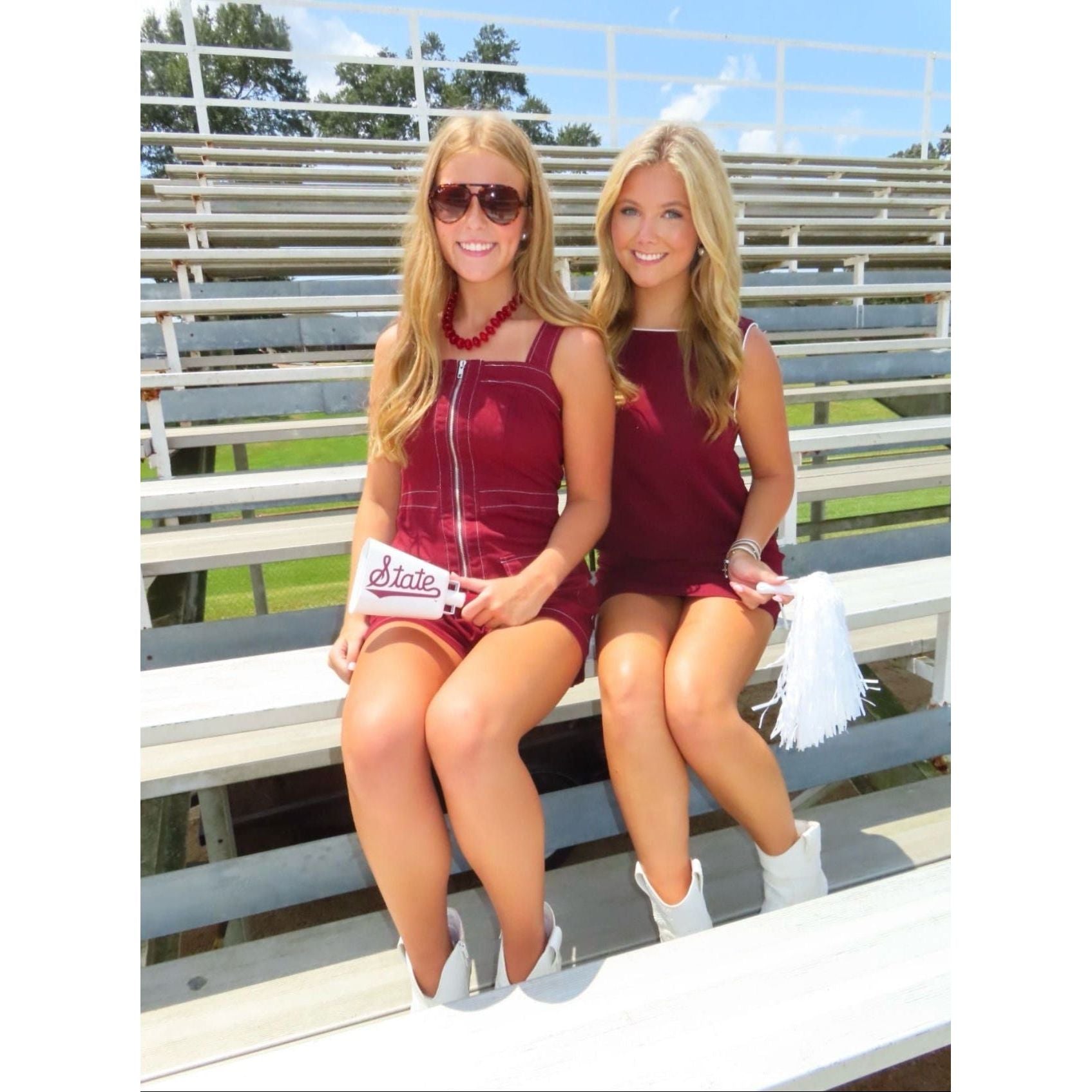 Two women sitting together on outdoor stadium bleachers, wearing matching sleeveless burgundy mini dresses and white cowboy boots. One holds a white 'Duke' clutch, and the other holds a white tassel.