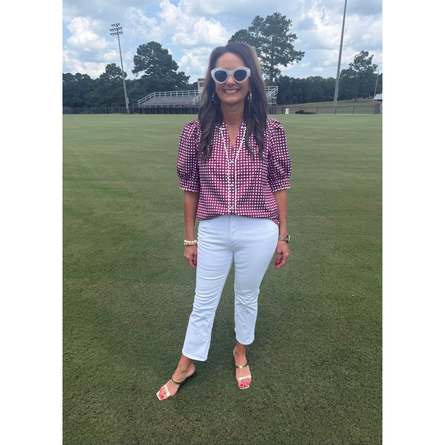 A woman posing on a green grass field, wearing a pink and white gingham short-sleeve button-up shirt, white pants, and pink sandals.