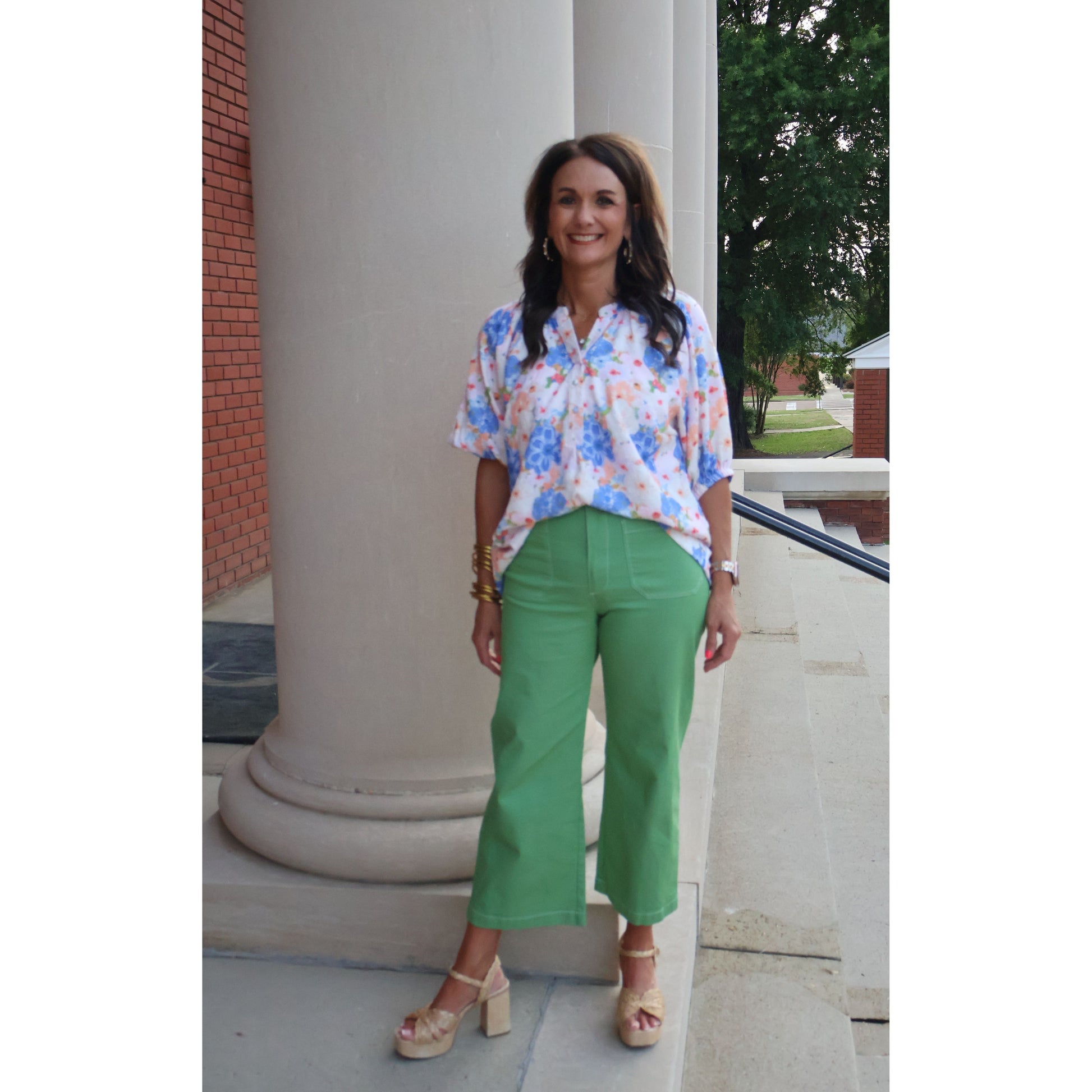 Woman in floral blouse and green pants standing outdoors near a columned building.
