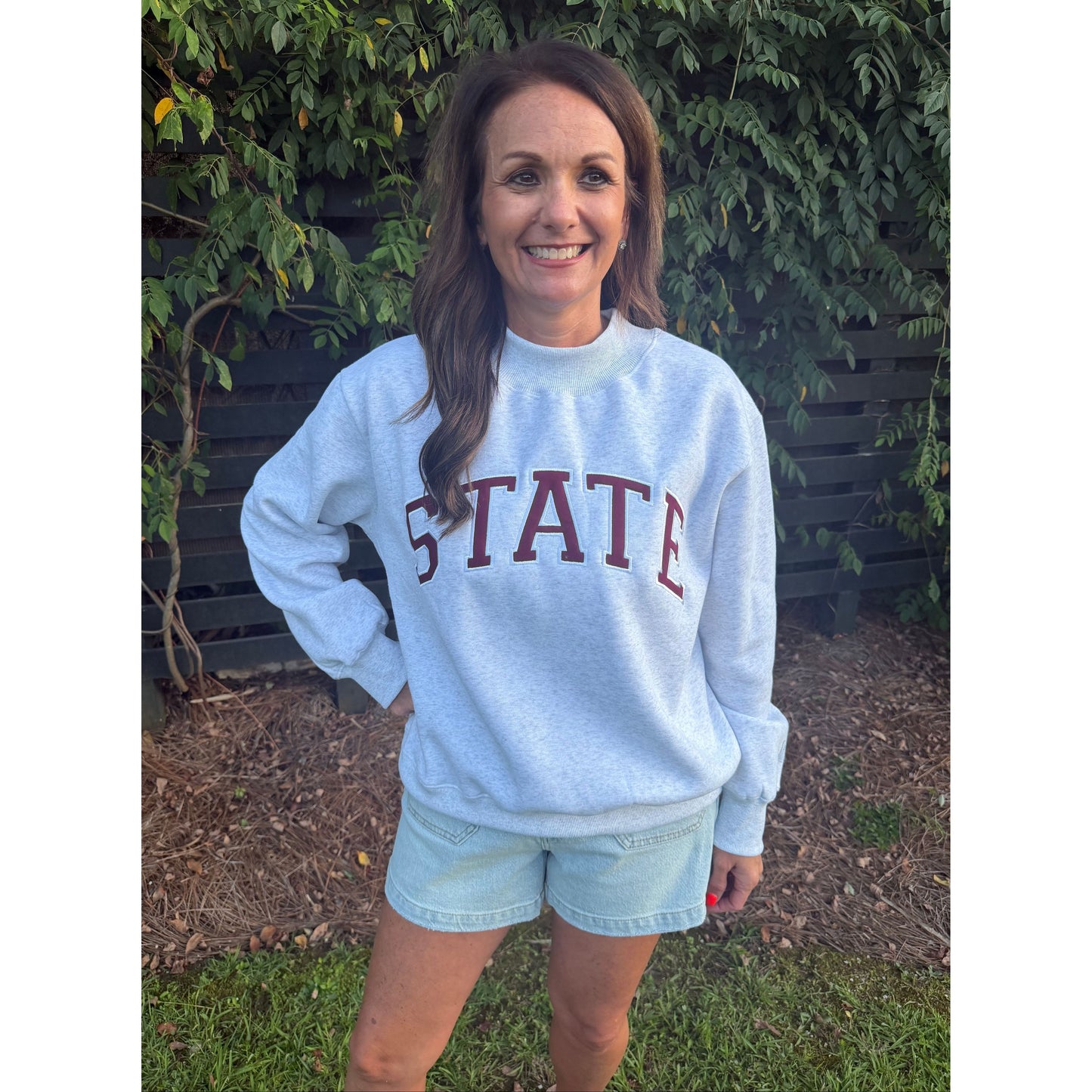 A woman wearing a light blue sweatshirt with 'STATE' printed in dark red letters, paired with light denim shorts, standing outdoors on a bench with greenery in the background.