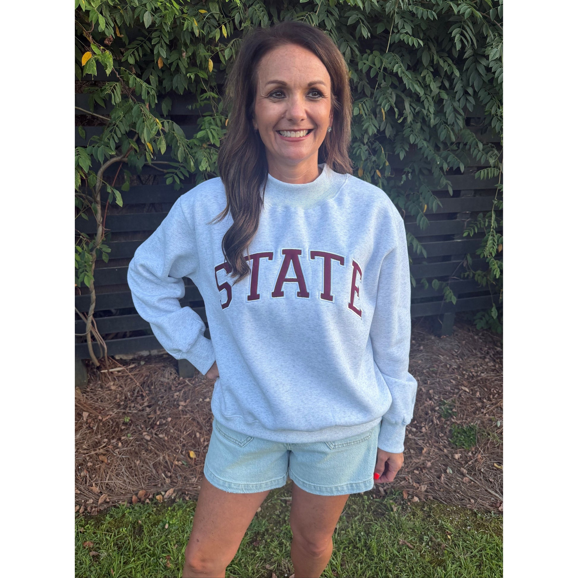 A woman wearing a light blue sweatshirt with 'STATE' printed in dark red letters, paired with light denim shorts, standing outdoors on a bench with greenery in the background.