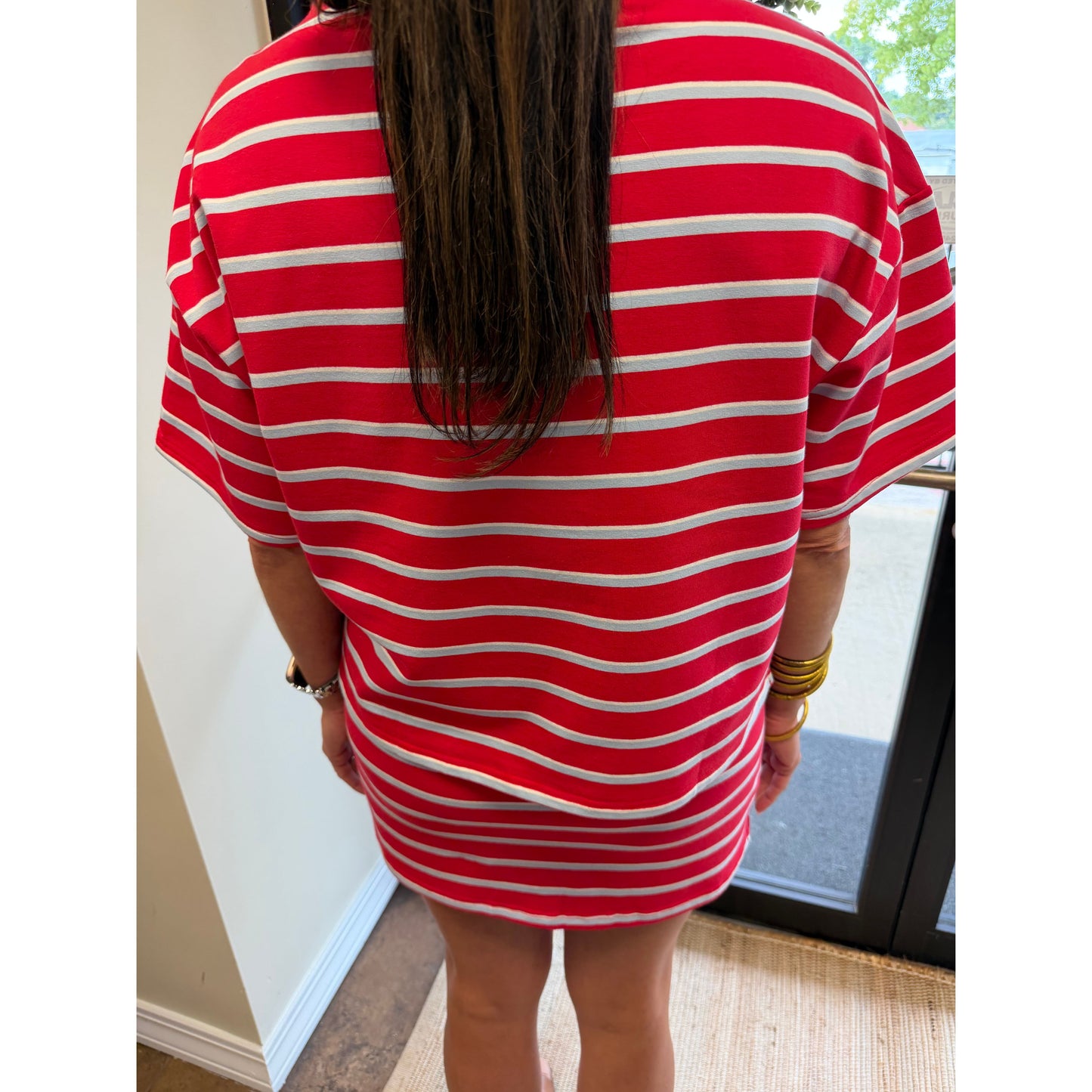 A person viewed from behind wearing an oversized red and white horizontally striped t-shirt dress, standing indoors near a glass door.