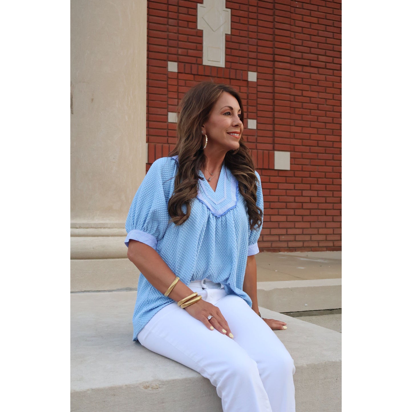 Woman in a light blue blouse and white pants standing against a brick wall.