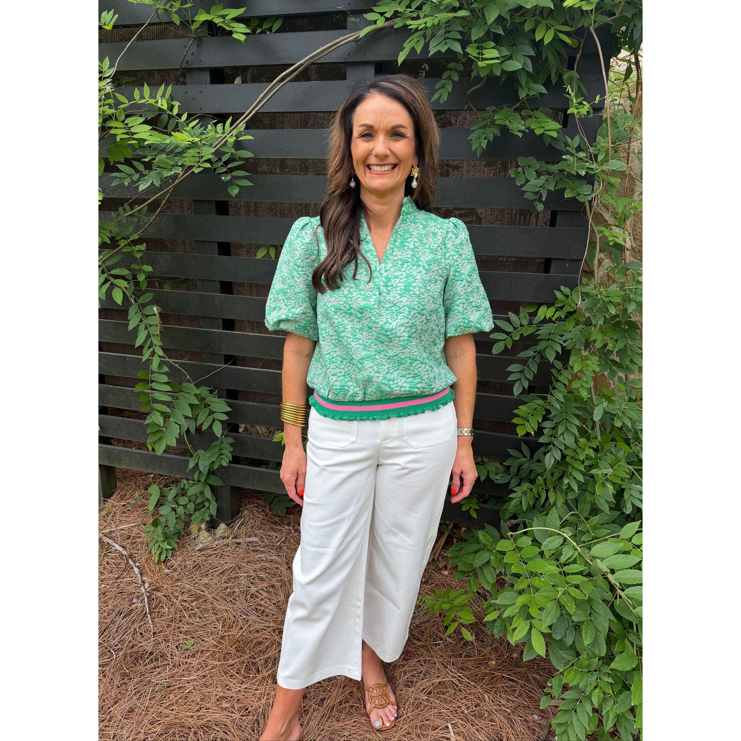 Woman in a green top and white pants standing outdoors with greenery around