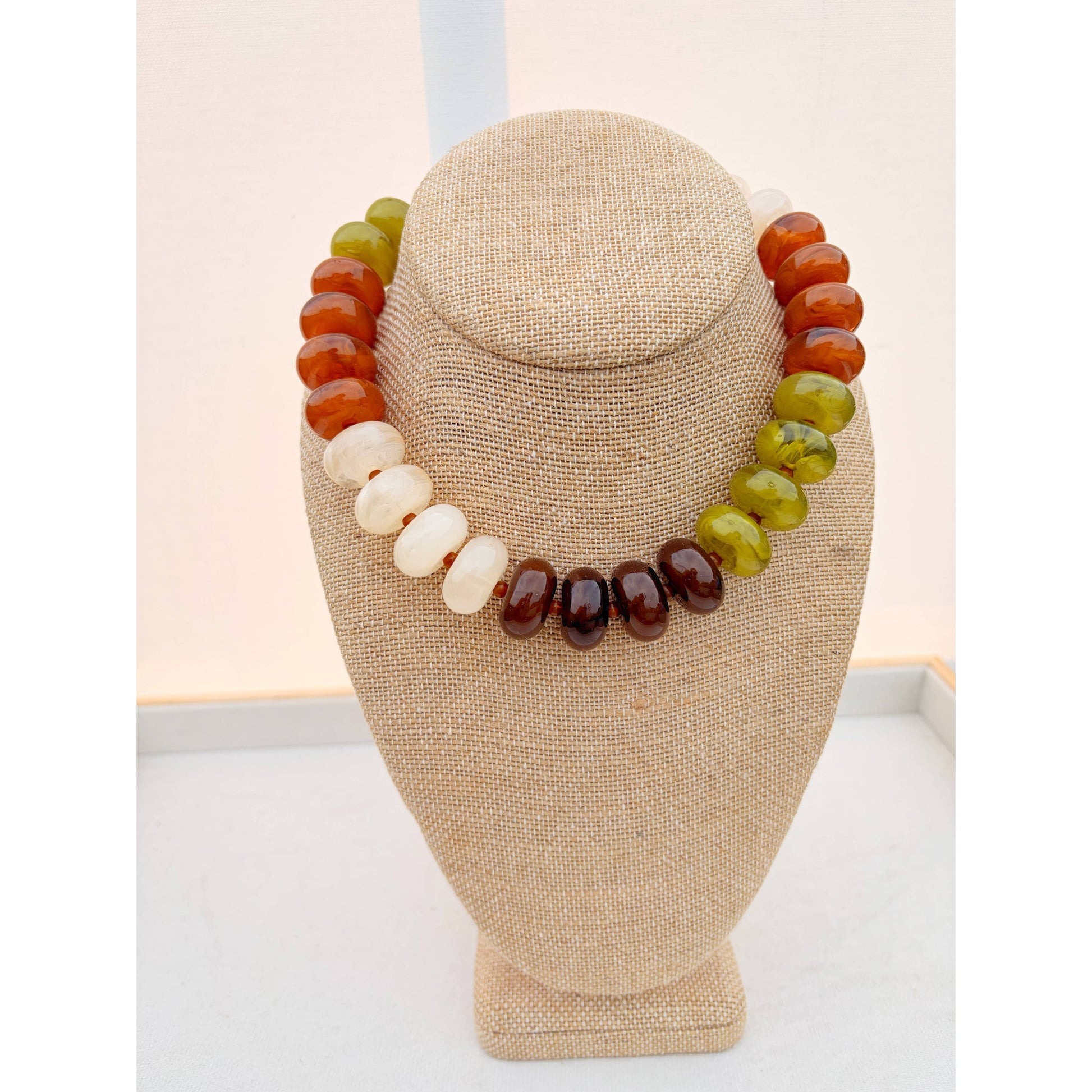 A multi-colored beaded necklace displayed on a beige fabric jewelry bust. The necklace features large, polished oval beads in shades of orange, olive green, cream white, and dark brown.