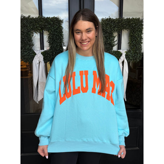 woman wearing a light blue sweatshirt with LULU MAC on it standing in front of black doors with wreaths.