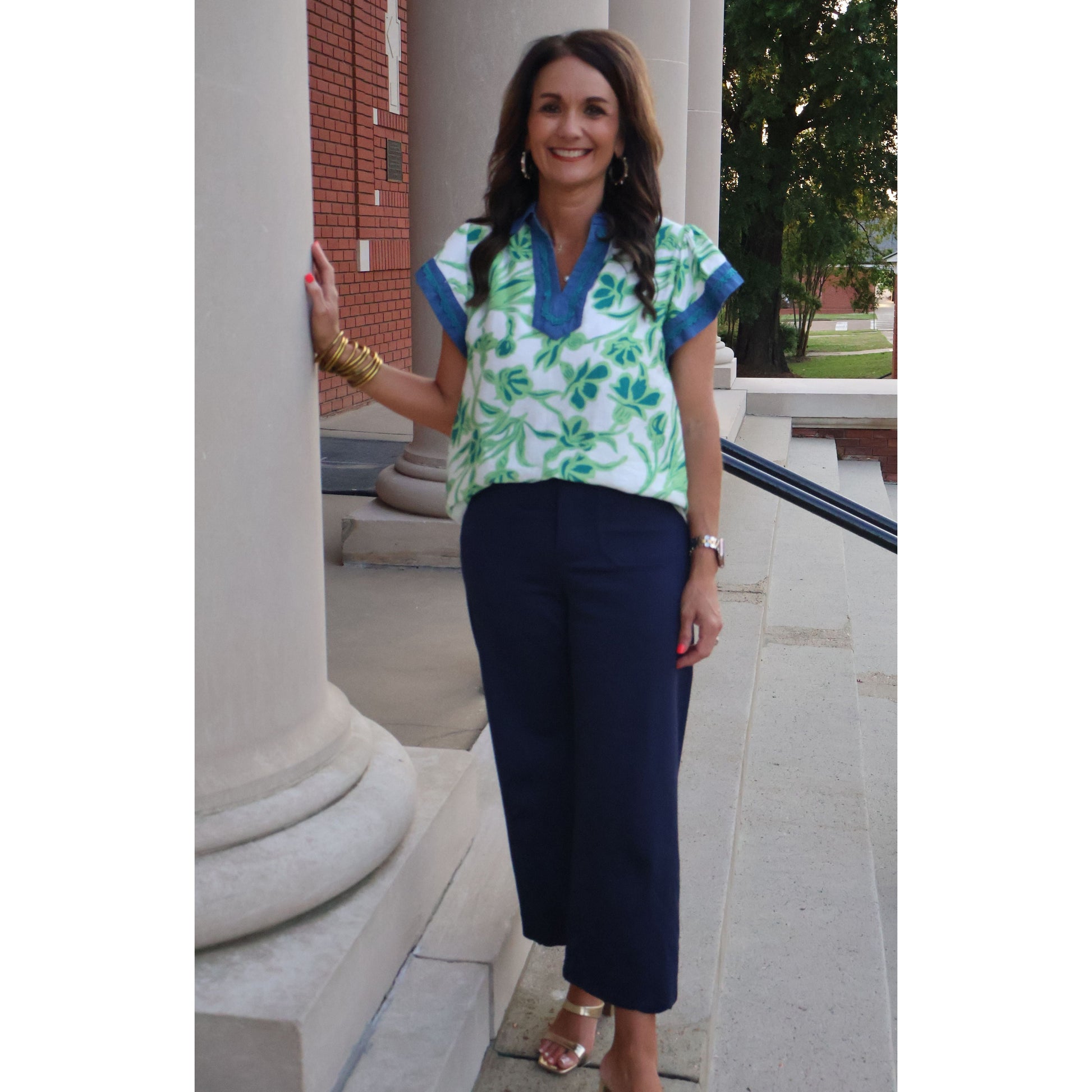 Woman in a floral top and blue pants standing outdoors near a building.
