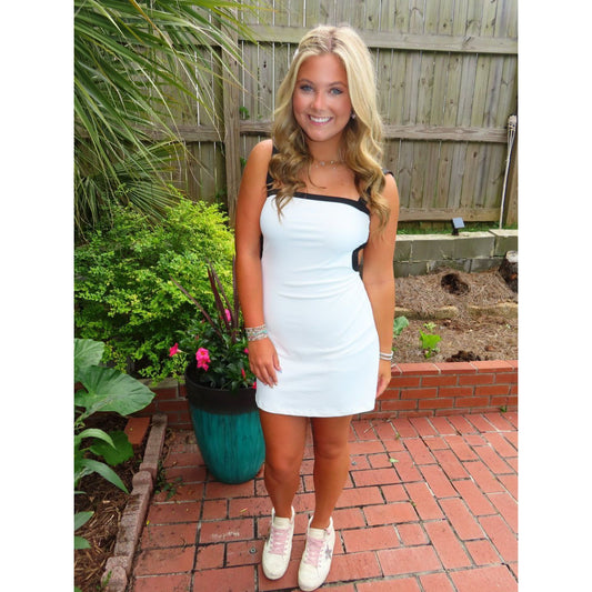Woman in a white dress standing outdoors on a brick patio with plants and a wooden fence in the background.