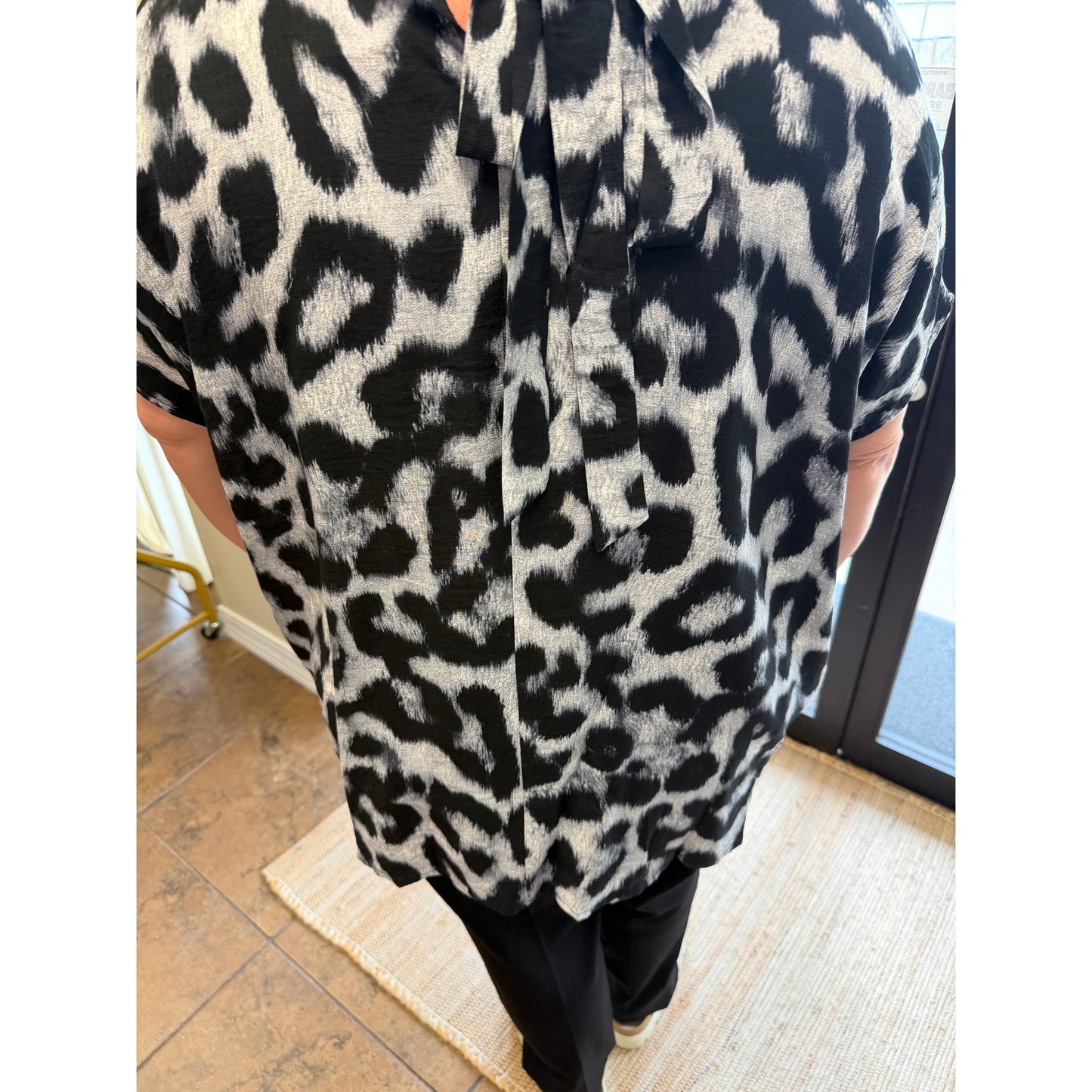 A person wearing a short-sleeve button-up shirt with a bold black and off-white leopard print pattern, paired with black pants, standing indoors on a woven rug.