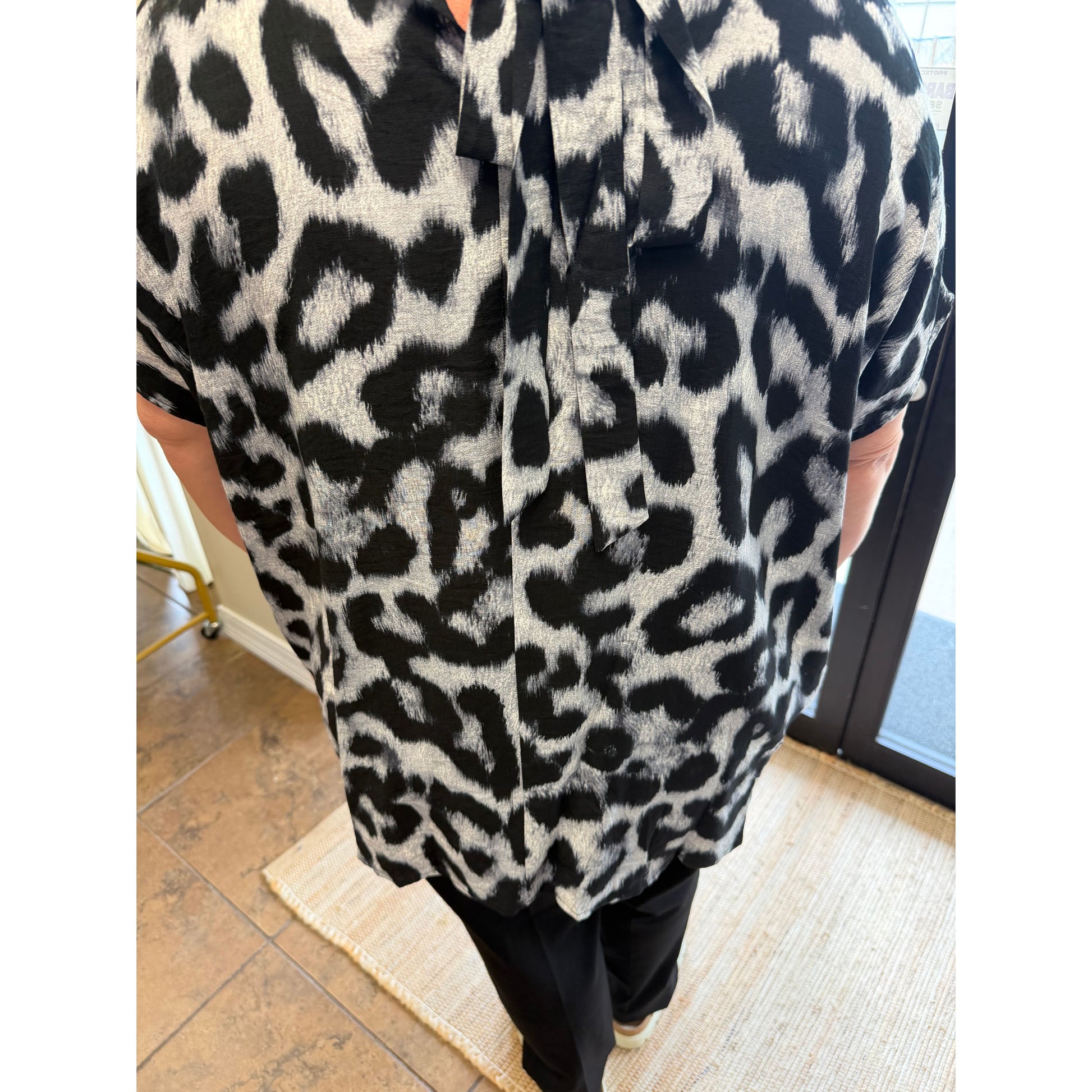 A person wearing a short-sleeve button-up shirt with a bold black and off-white leopard print pattern, paired with black pants, standing indoors on a woven rug.