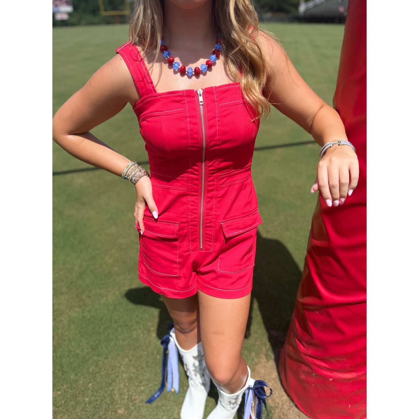 A woman wearing a bright red sleeveless romper with a front zipper and flap pockets, standing outdoors on a grassy field next to a red structure.