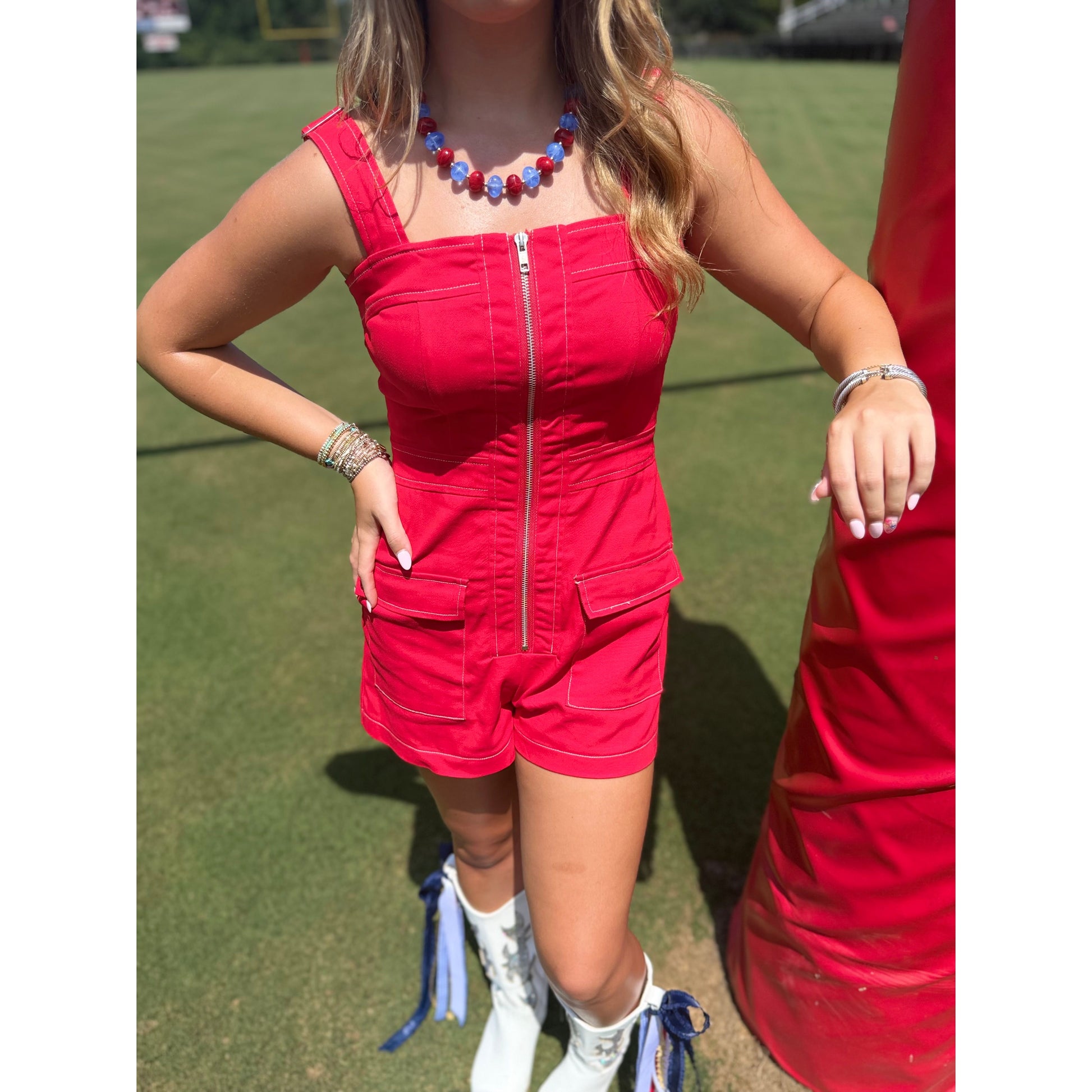 A woman wearing a bright red sleeveless romper with a front zipper and flap pockets, standing outdoors on a grassy field next to a red structure.