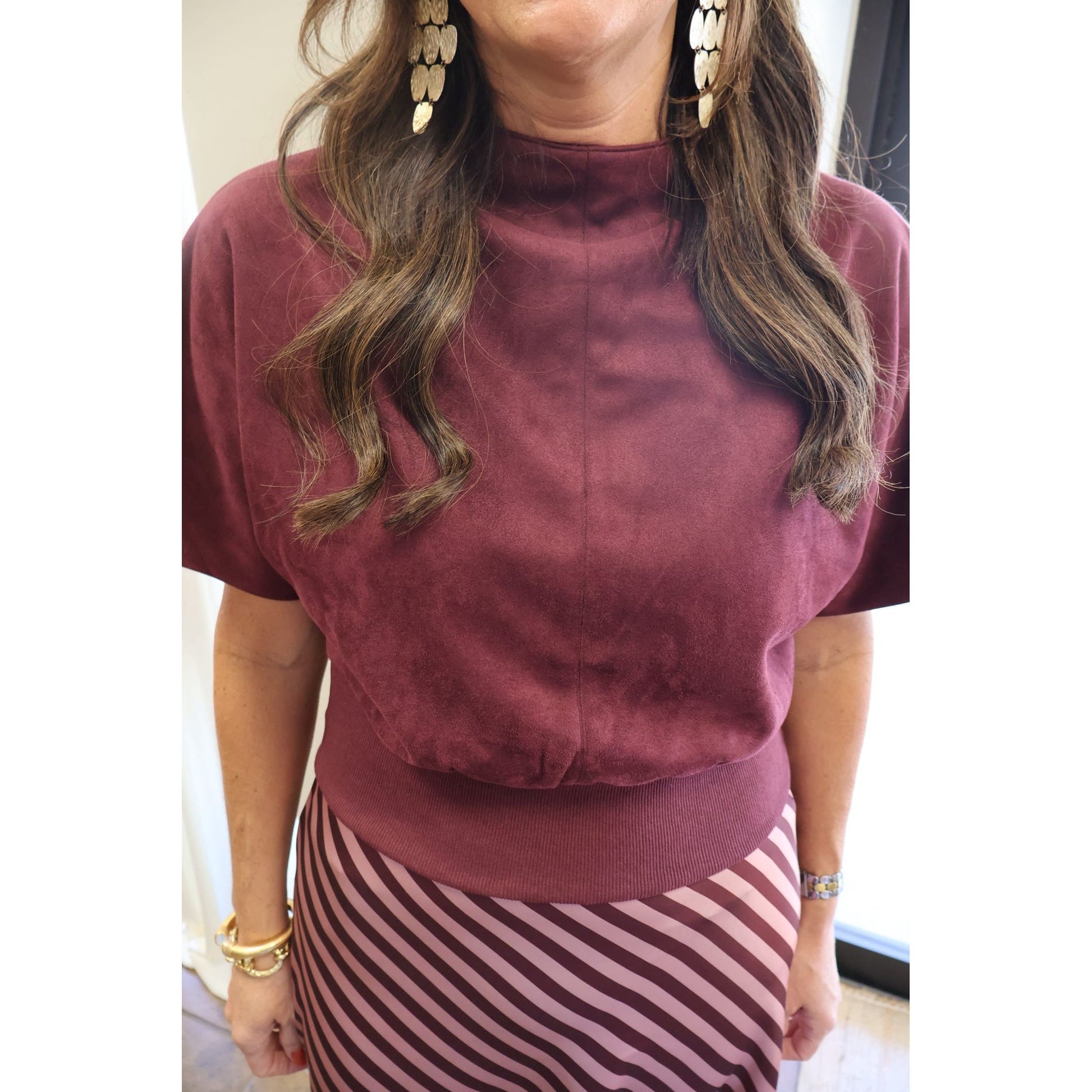 A woman wearing a short-sleeve mock neck top in a dusty burgundy or plum velvet-like fabric, paired with a pink and burgundy striped skirt. She is wearing large gold disc earrings and bracelets.