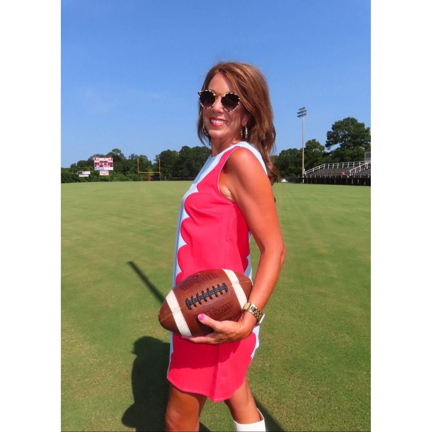 A woman wearing a bright pink and white sleeveless dress stands on a green football field holding a brown football under her arm. She is wearing gold-framed sunglasses, and the background shows a blue sky, trees, and stadium bleachers.