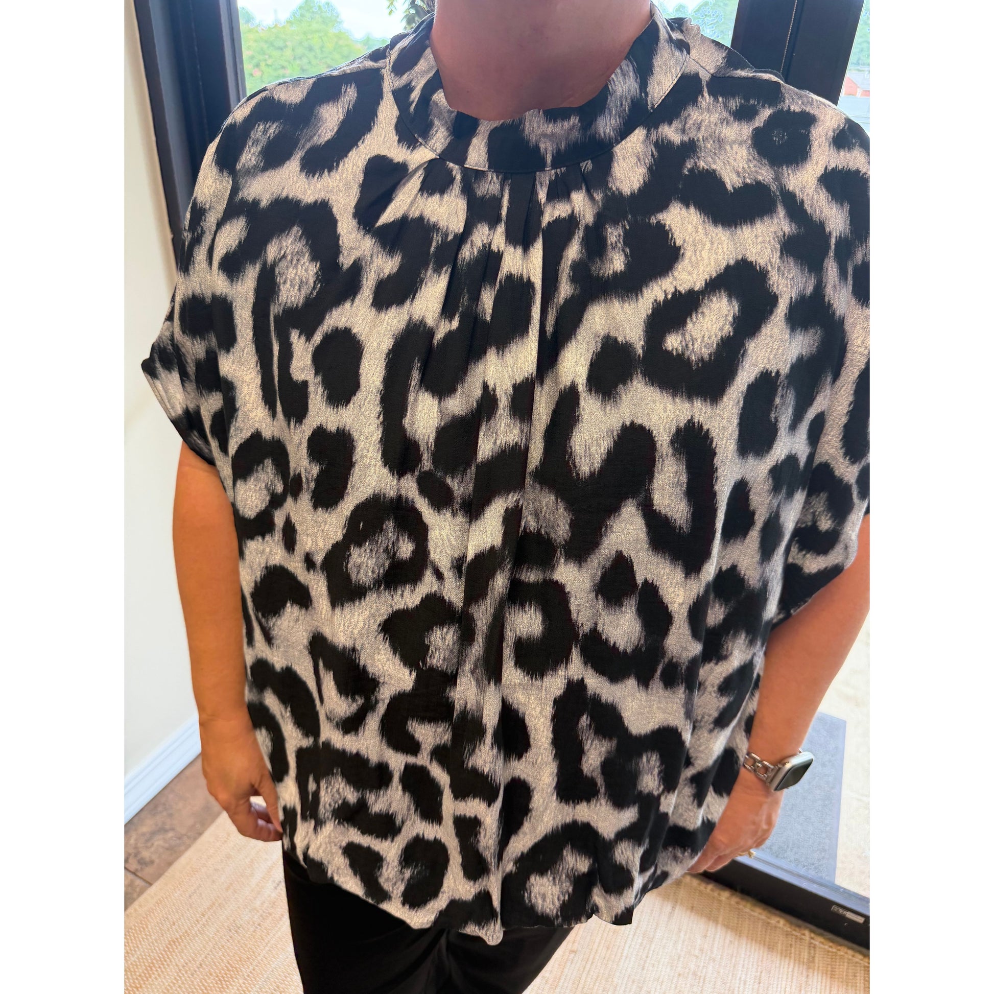 A person wearing a short-sleeve leopard print blouse with a high gathered neckline, paired with black pants, standing indoors.