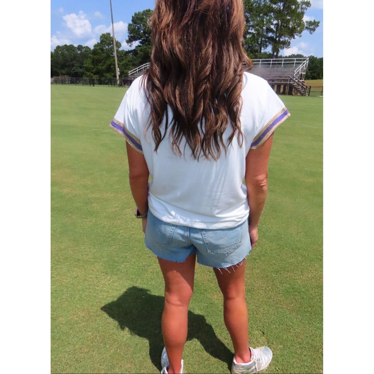 A woman viewed from behind wearing a white short-sleeve t-shirt with purple and blue striped trim on the sleeves, paired with light blue denim shorts, standing on a green grass field.