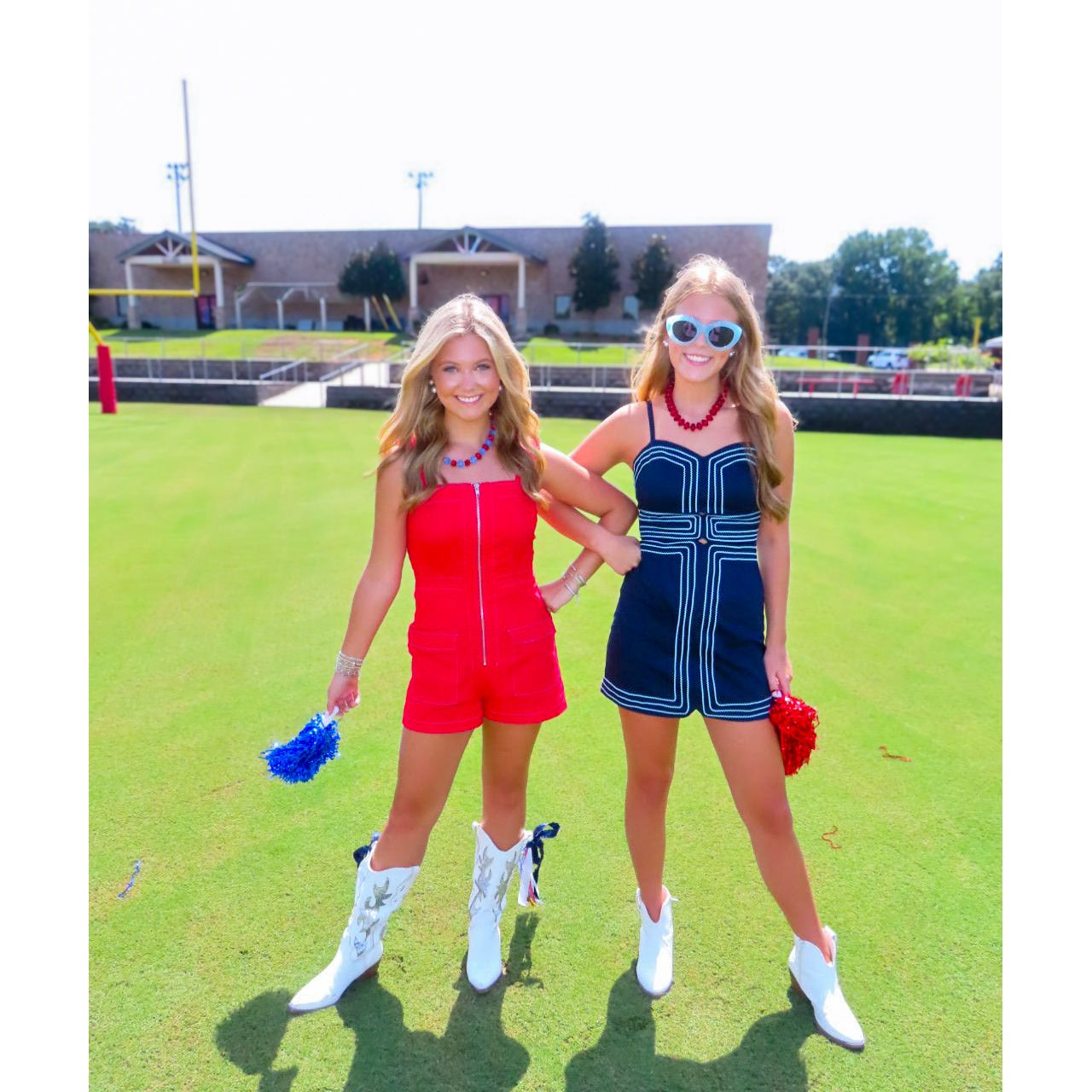 Two women posing on a green grass field wearing matching red and navy blue romper outfits with white cowboy boots.