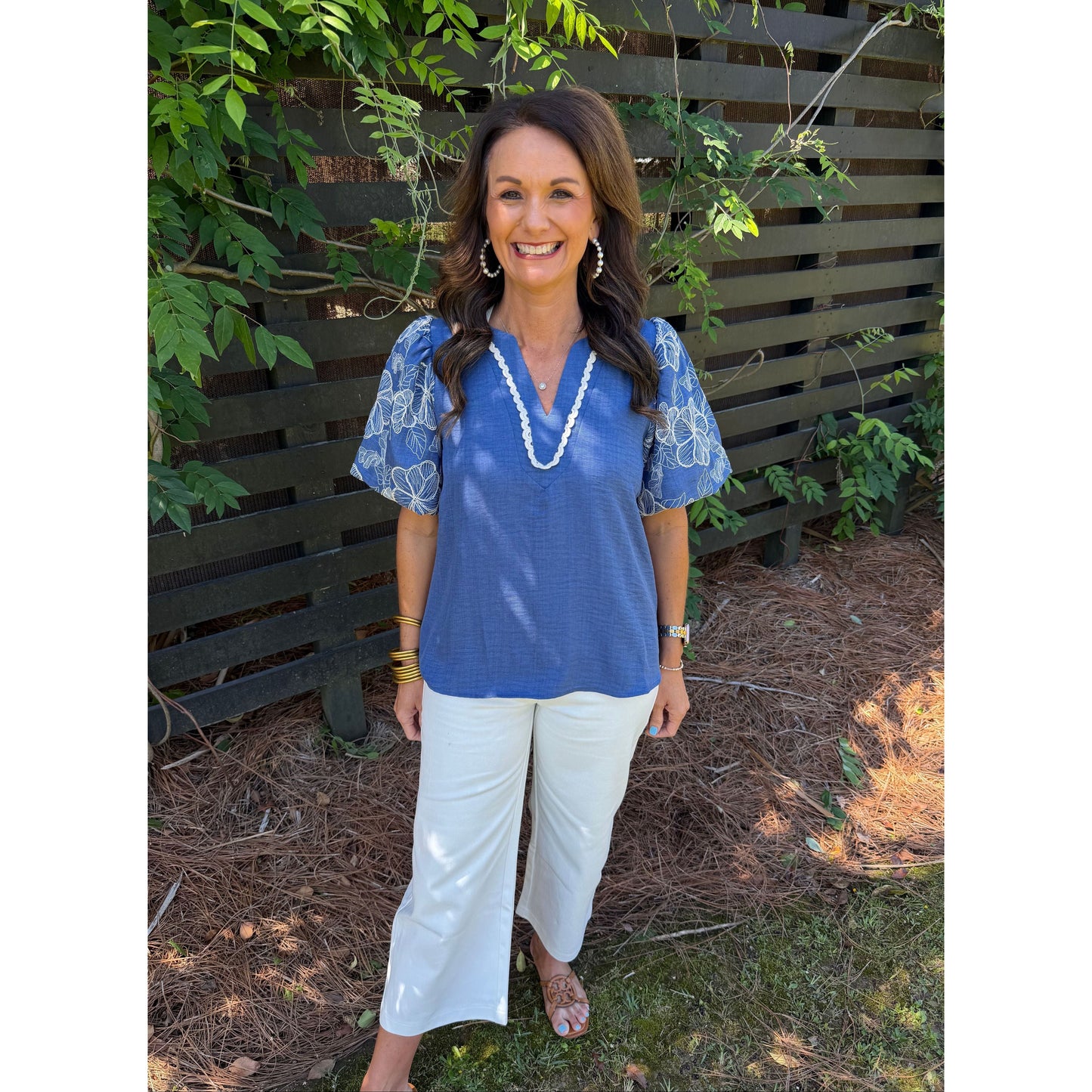 Woman in a blue top and white pants standing outdoors with greenery around