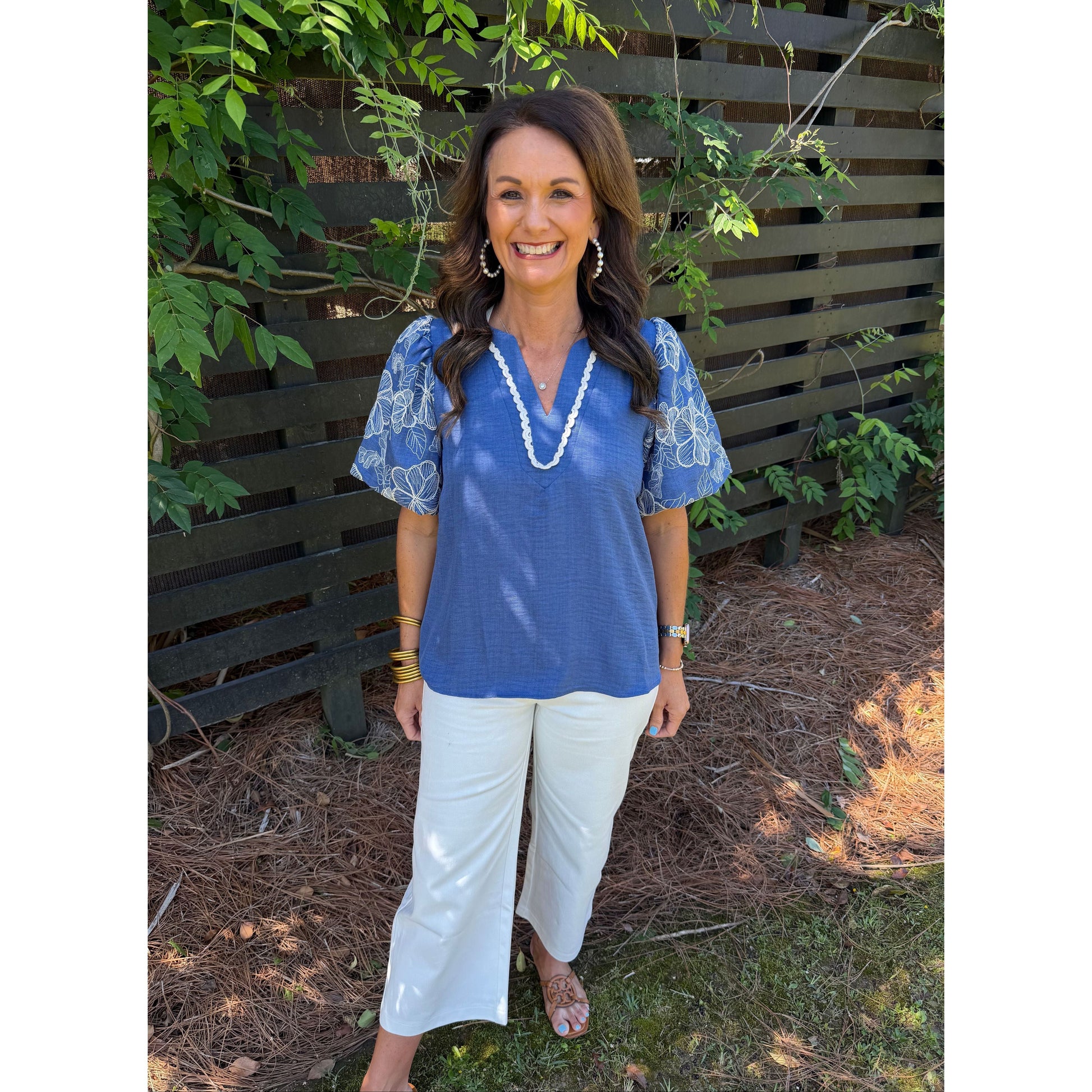 Woman in a blue top and white pants standing outdoors with greenery around