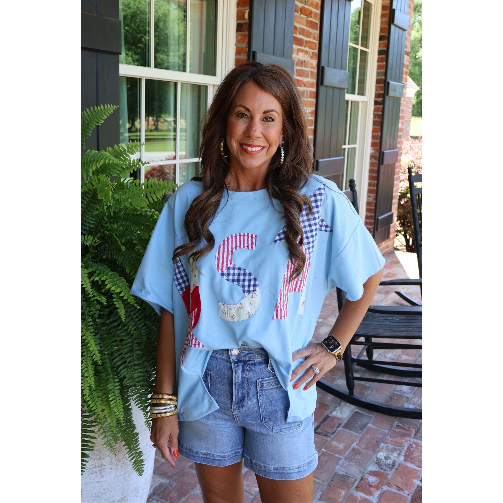 Woman wearing a light blue t-shirt with graphic designs and denim shorts standing on a patio.