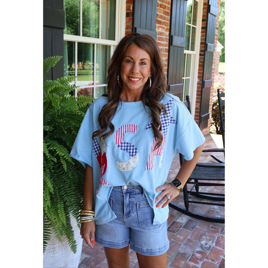 Woman wearing a light blue t-shirt with graphic designs and denim shorts standing on a patio.
