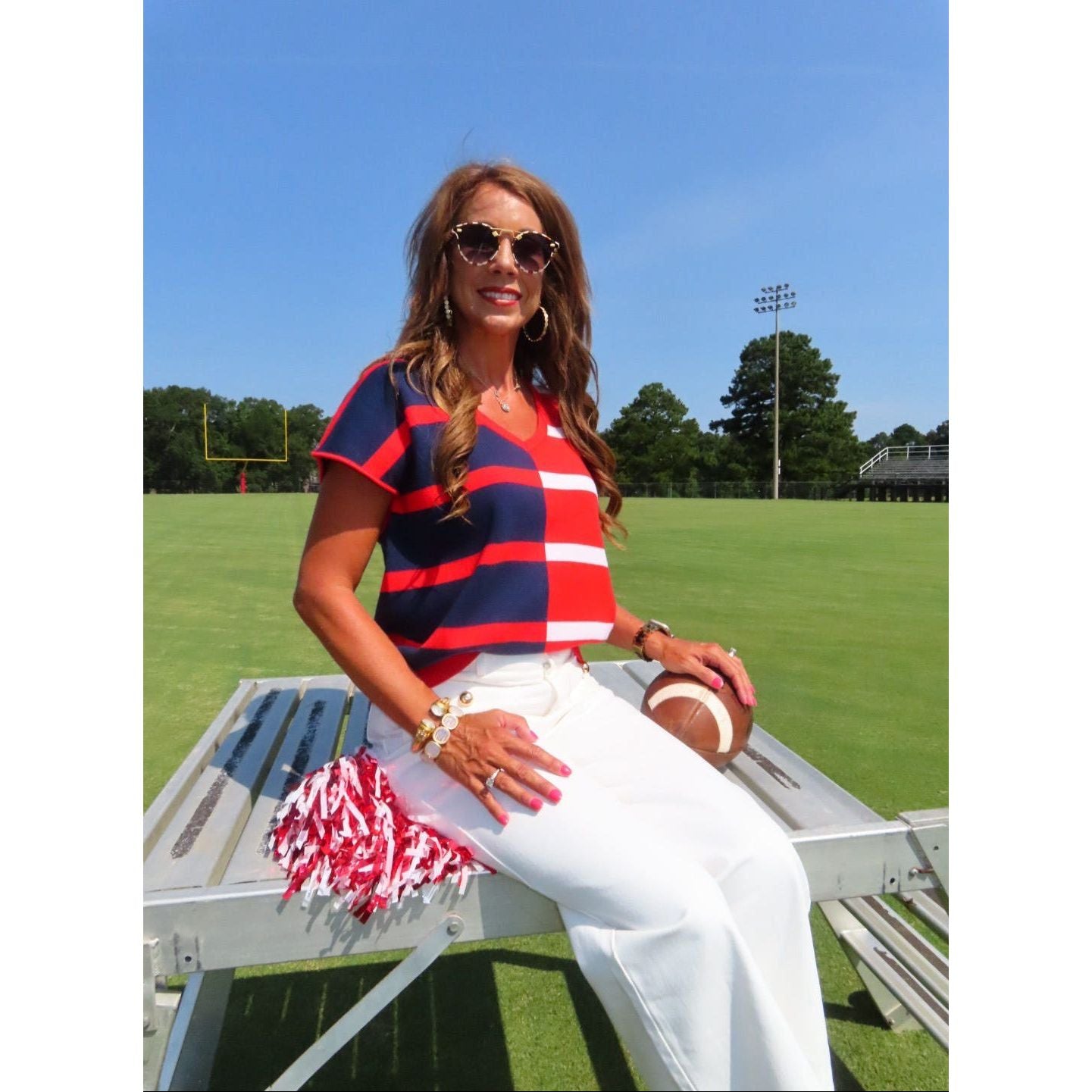 A woman wearing a red, navy, and white striped short-sleeve V-neck top, white pants, and sunglasses sits on a metal bleachers at a sunny outdoor sports field.