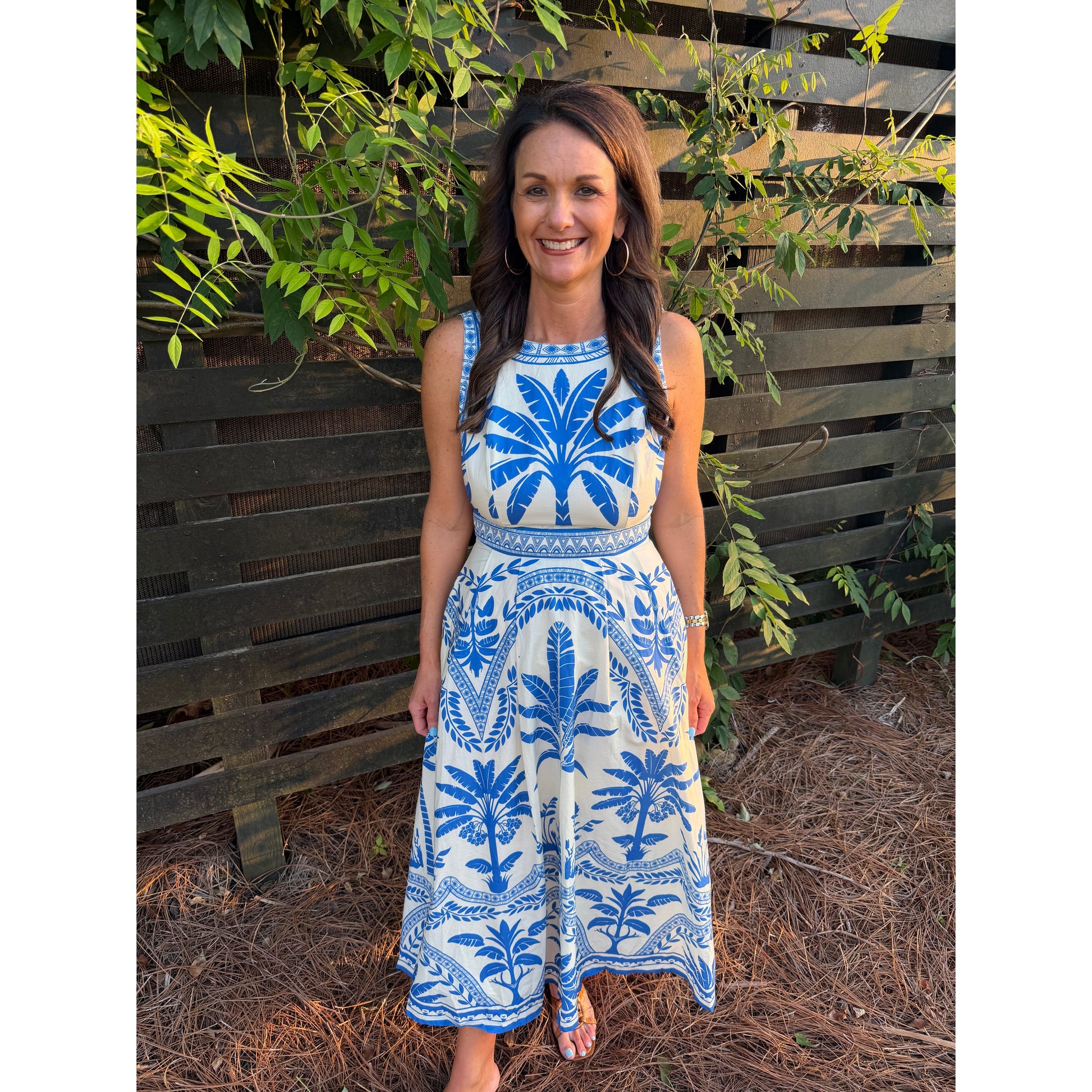 Woman wearing a blue and white patterned dress standing in front of a wooden fence with greenery.