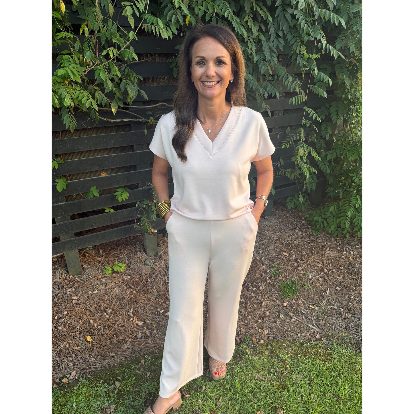 A woman wearing a matching cream short-sleeve V-neck top and wide-leg trousers stands outdoors on grass in front of a dark wooden fence and green foliage.