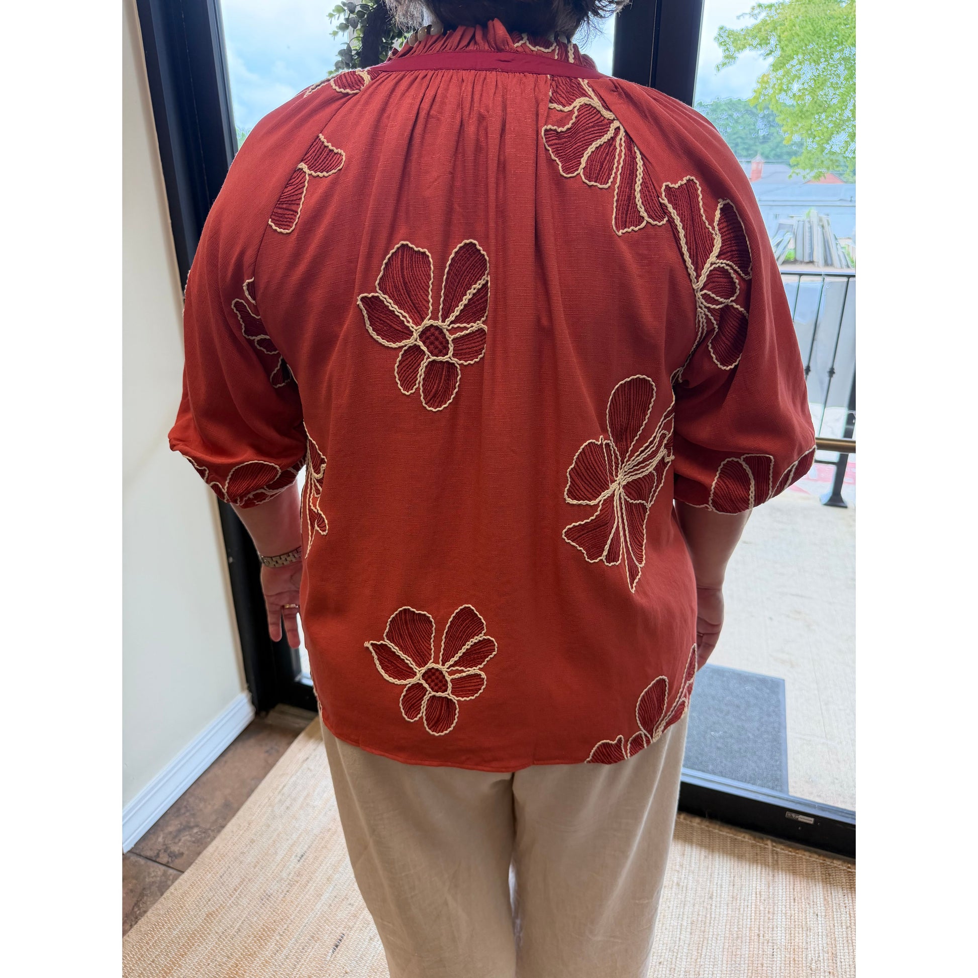 A person viewed from behind wearing a rust-orange short-sleeve blouse with large cream floral embroidery, paired with beige pants, standing indoors near a glass door.