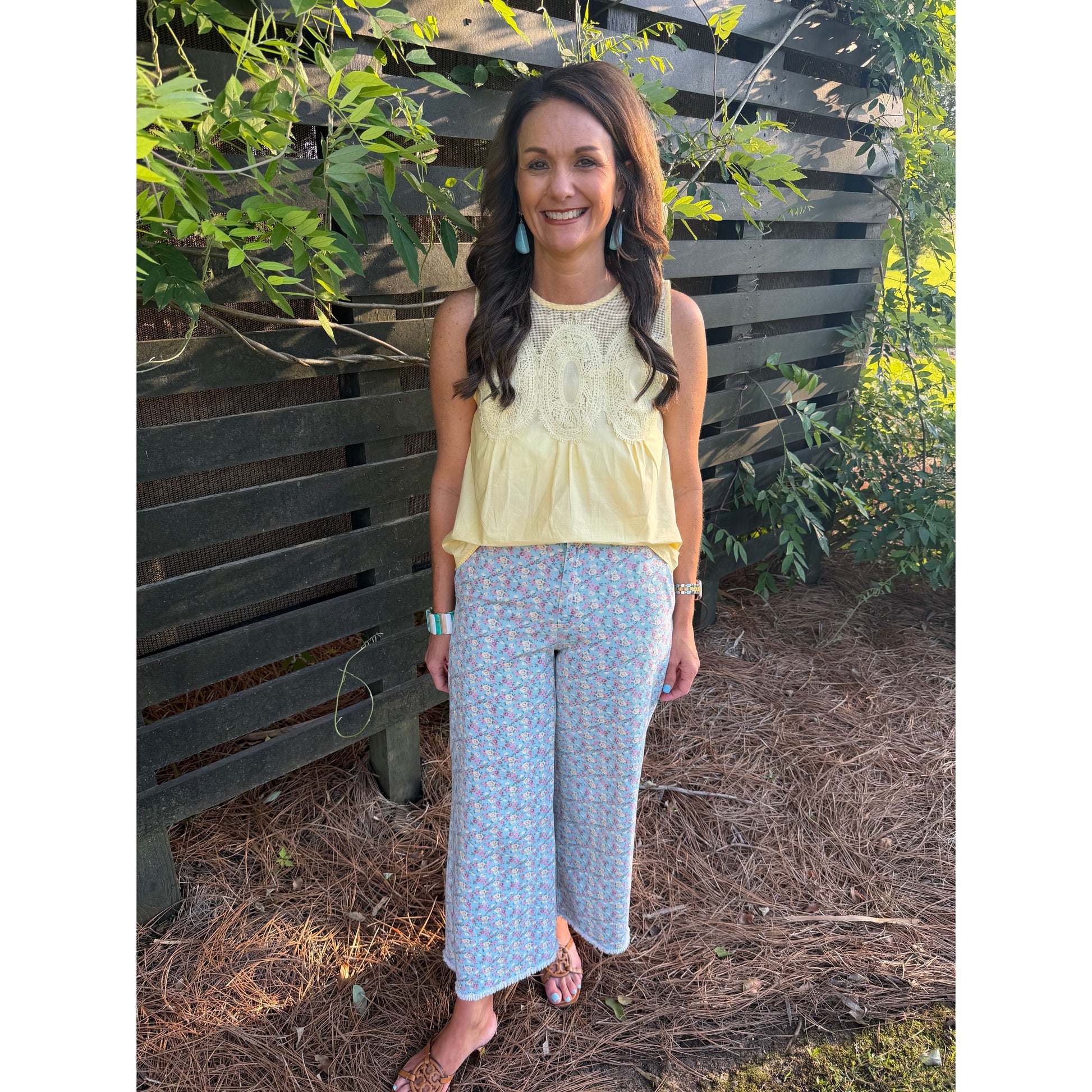 Woman wearing a yellow top and patterned pants standing outdoors with greenery in the background