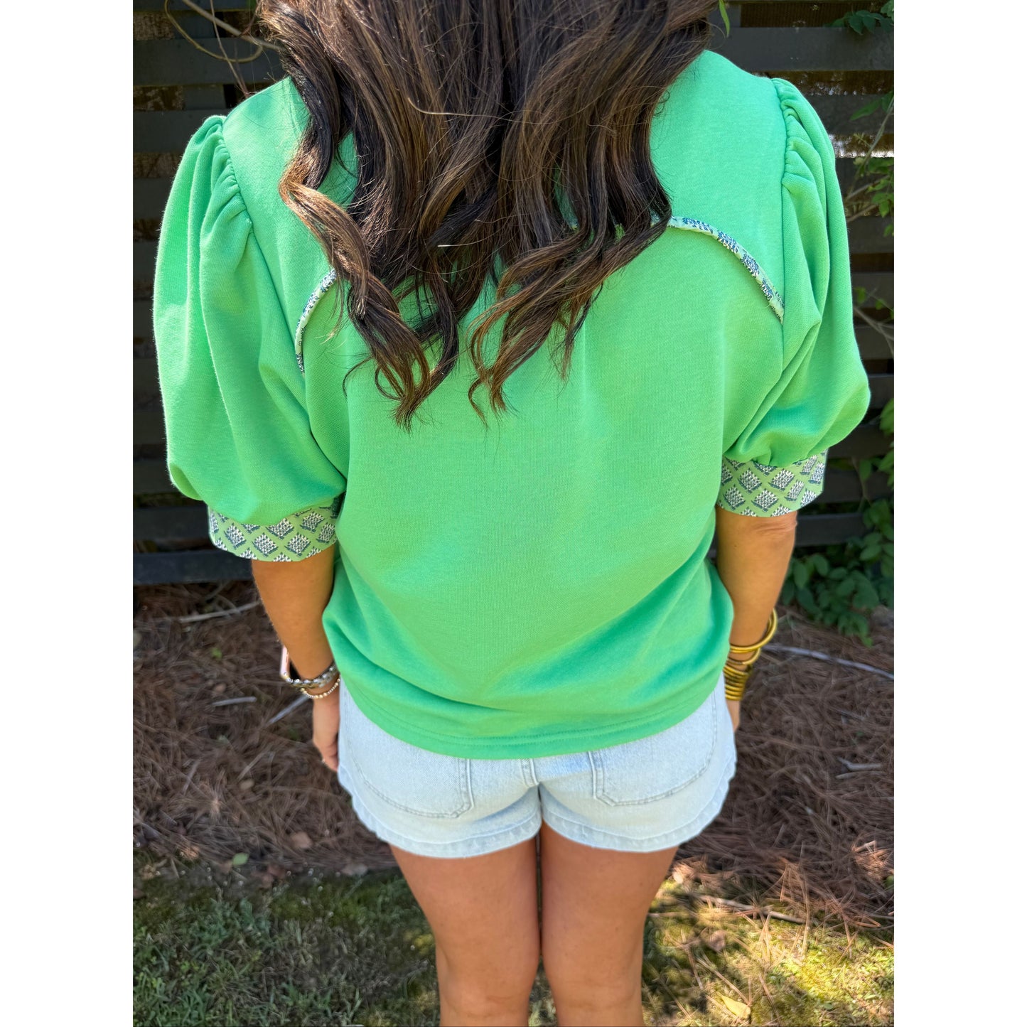 Person wearing a green shirt with patterned trim and white shorts, standing outdoors.