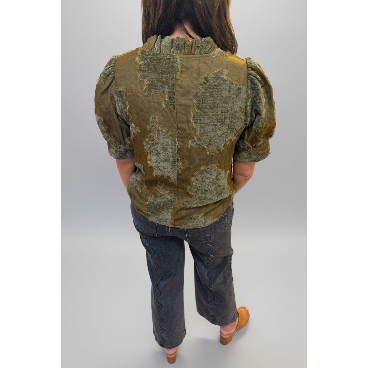 A person viewed from behind wearing a short-sleeve blouse with a distressed patchwork pattern in olive green and brown tones, paired with dark gray pants and tan shoes, standing in an indoor room with tiled floors and a white column.