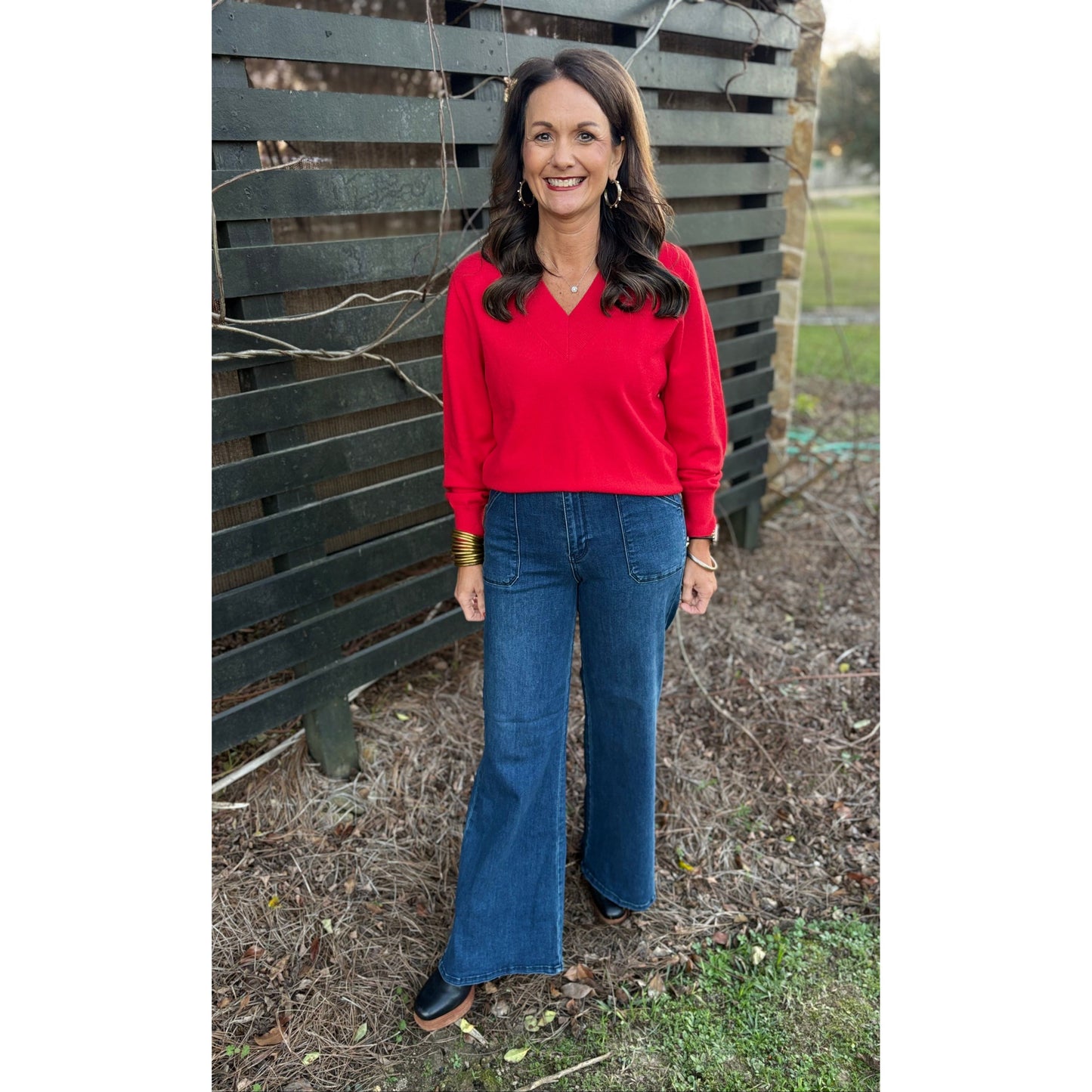 Woman wearing a red sweater and blue jeans standing in front of a wooden wall.