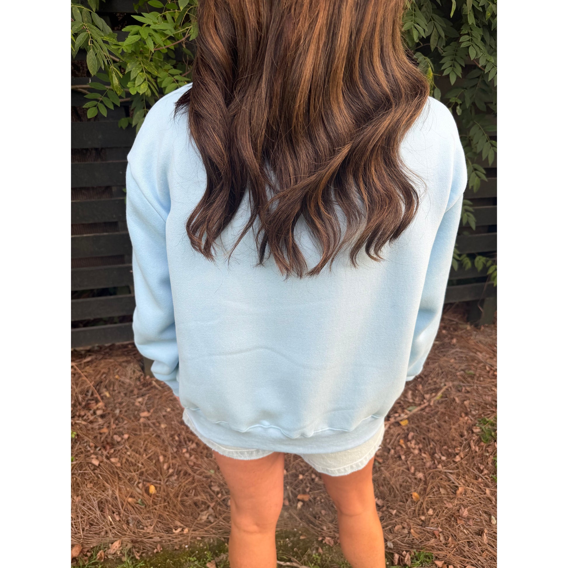 A person viewed from behind wearing a light blue sweatshirt and white denim shorts, standing outdoors on a mulch ground with green foliage and a dark fence in the background.