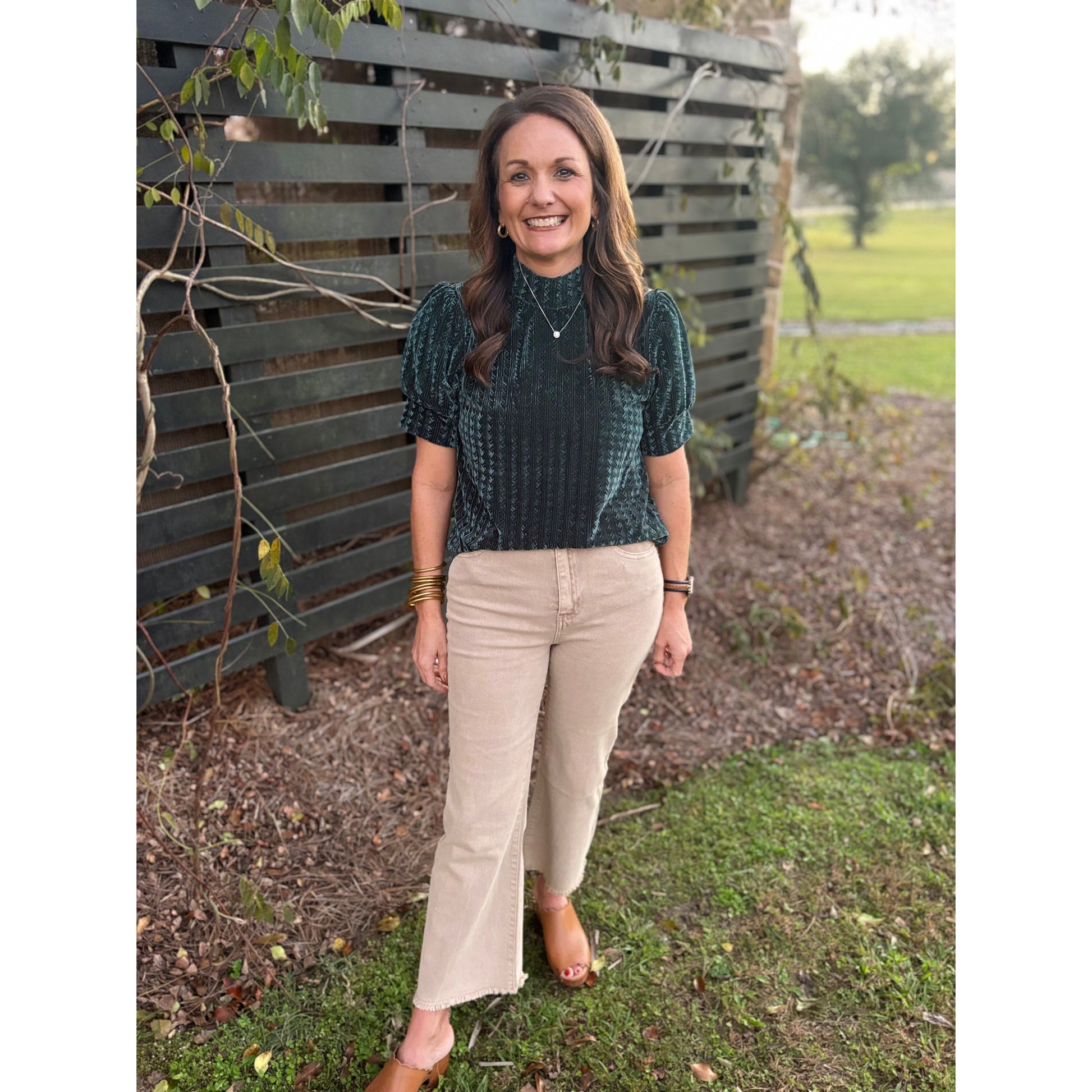 Woman standing outdoors wearing a green patterned top and beige pants.