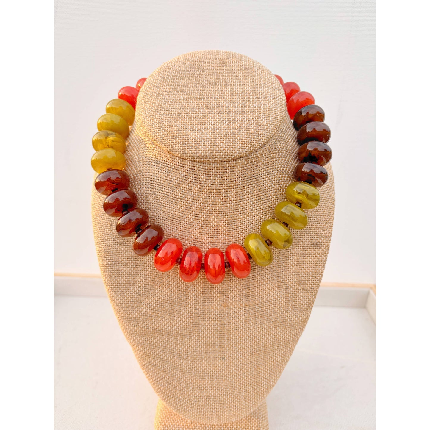 A beaded necklace made of polished oval beads in a gradient of yellow-green, orange-red, and dark brown, displayed on a beige fabric-covered jewelry bust against a white background.