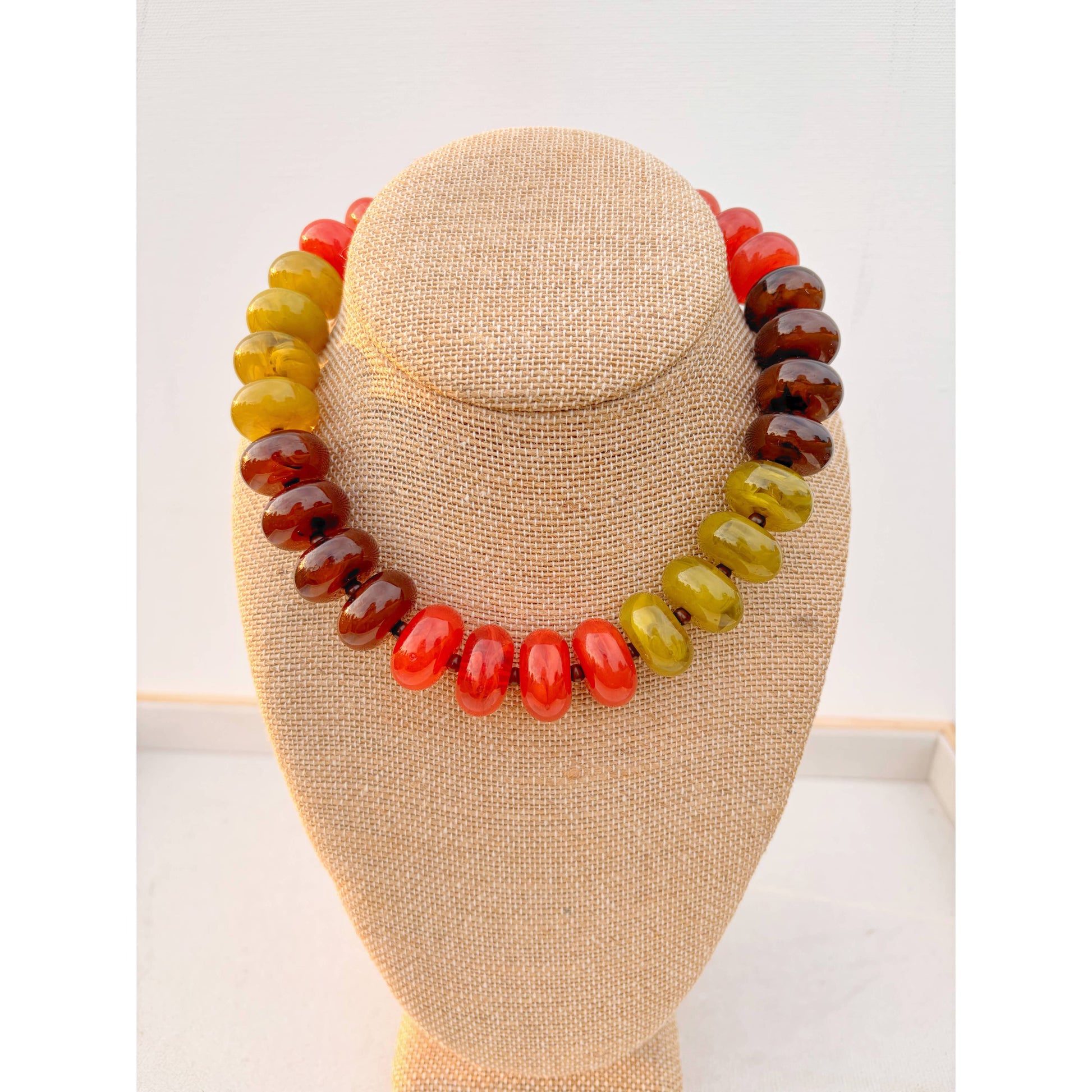A beaded necklace made of polished oval beads in a gradient of yellow-green, orange-red, and dark brown, displayed on a beige fabric-covered jewelry bust against a white background.