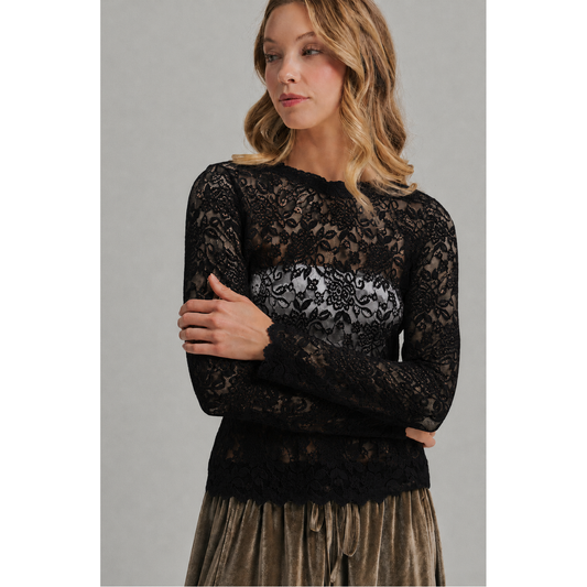 A woman wearing a black lace long-sleeve top with a sheer floral pattern over a gray underlayer, paired with a bronze velvet skirt, against a neutral gray background.