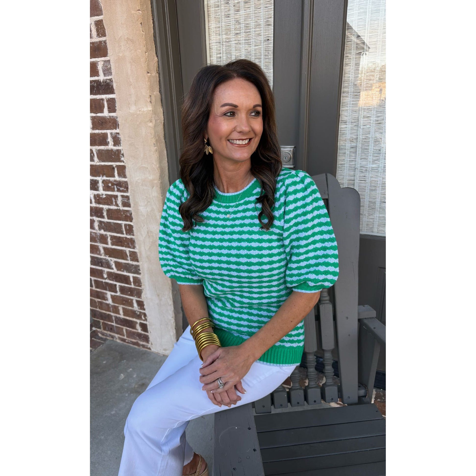 Woman wearing a green and white striped sweater and white pants sitting on a chair.