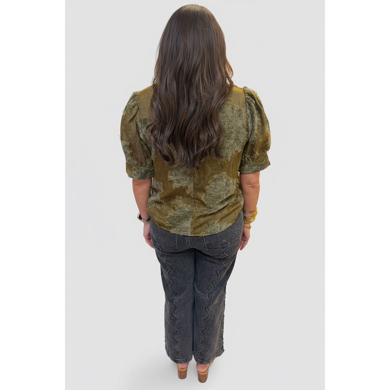 A woman viewed from behind wearing a short-sleeve olive green and brown patterned blouse with dark gray pants, standing in a tiled indoor hallway.