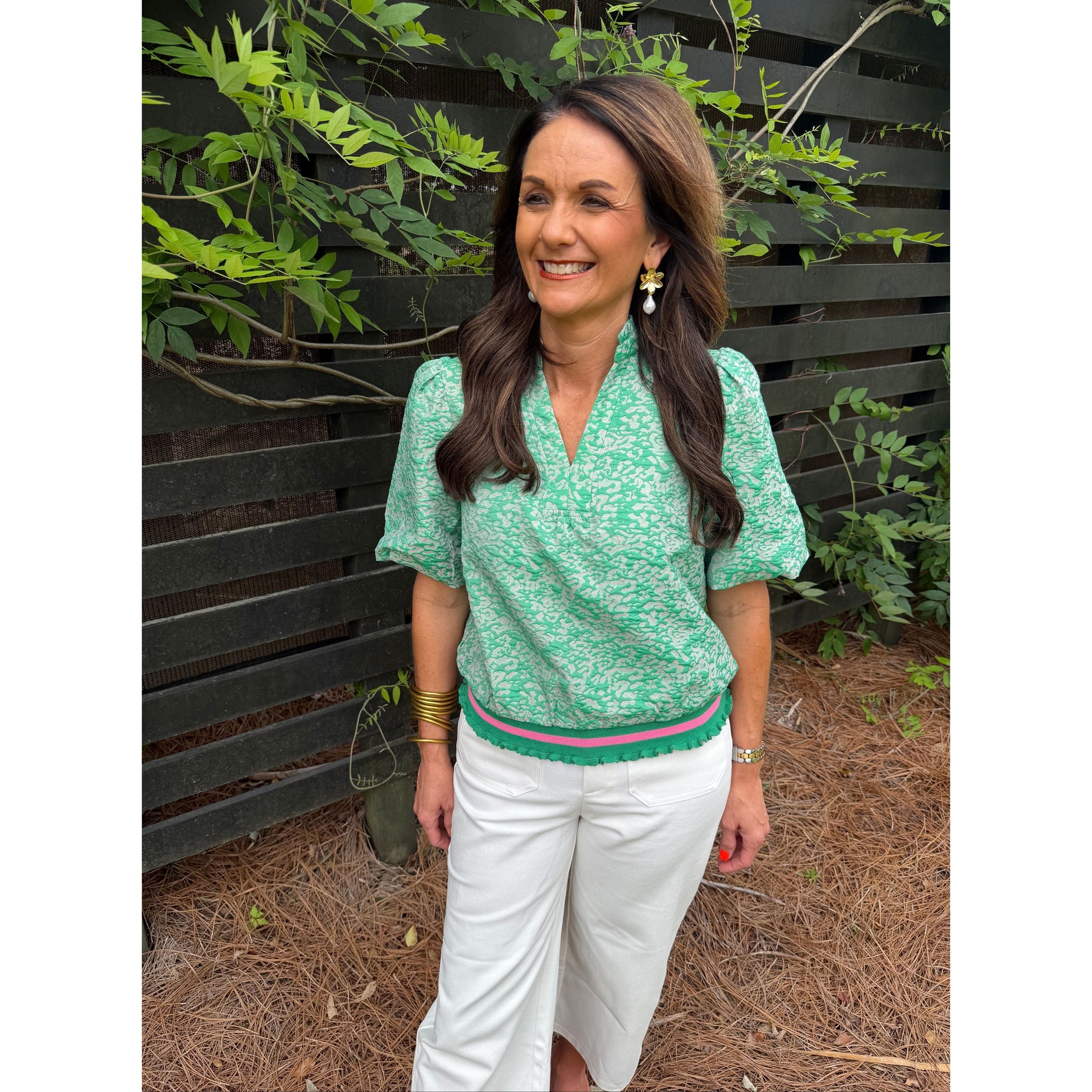 Woman in a green patterned top and white pants lying on a bench with plants around.