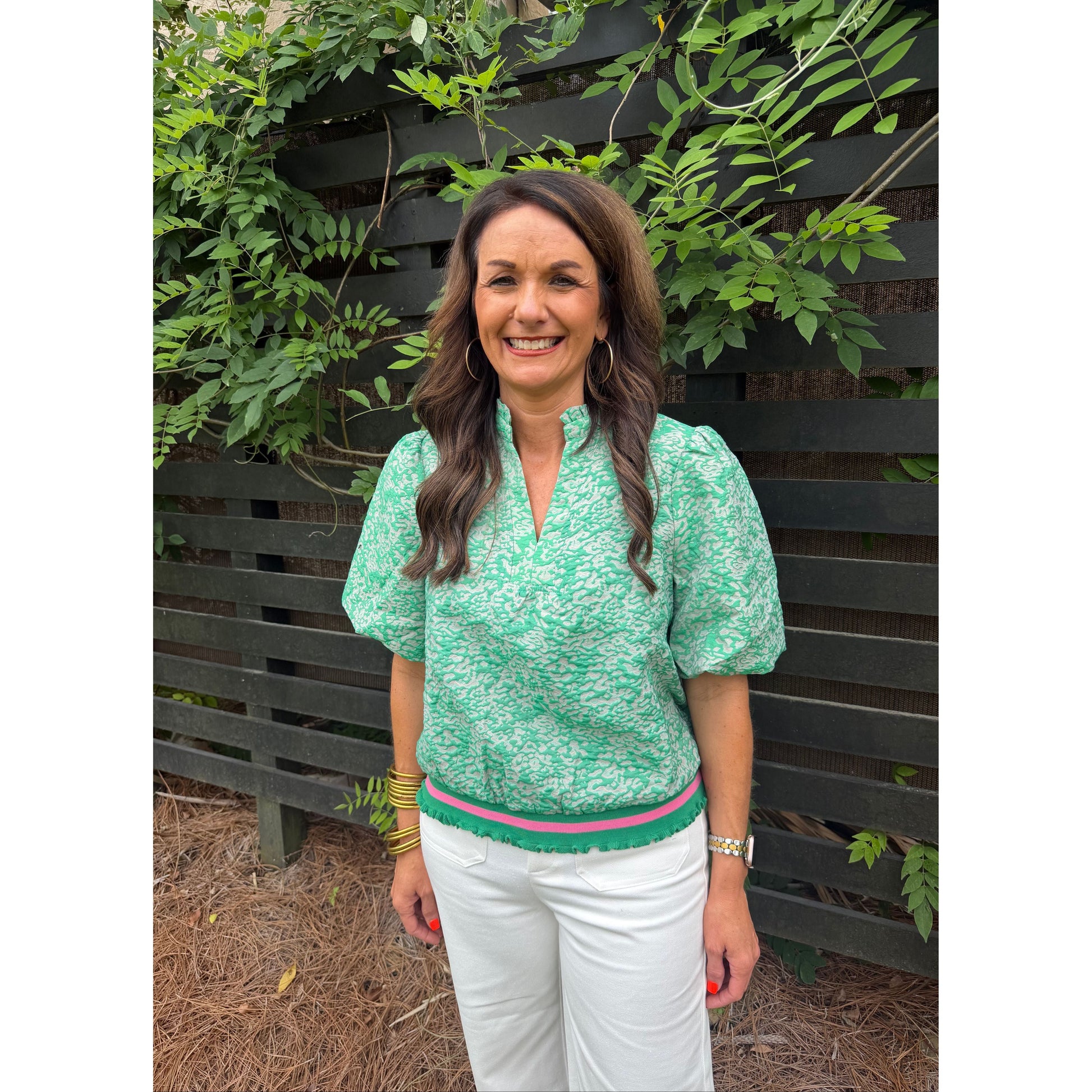 Woman wearing a green patterned top and white pants standing outdoors with plants in the background