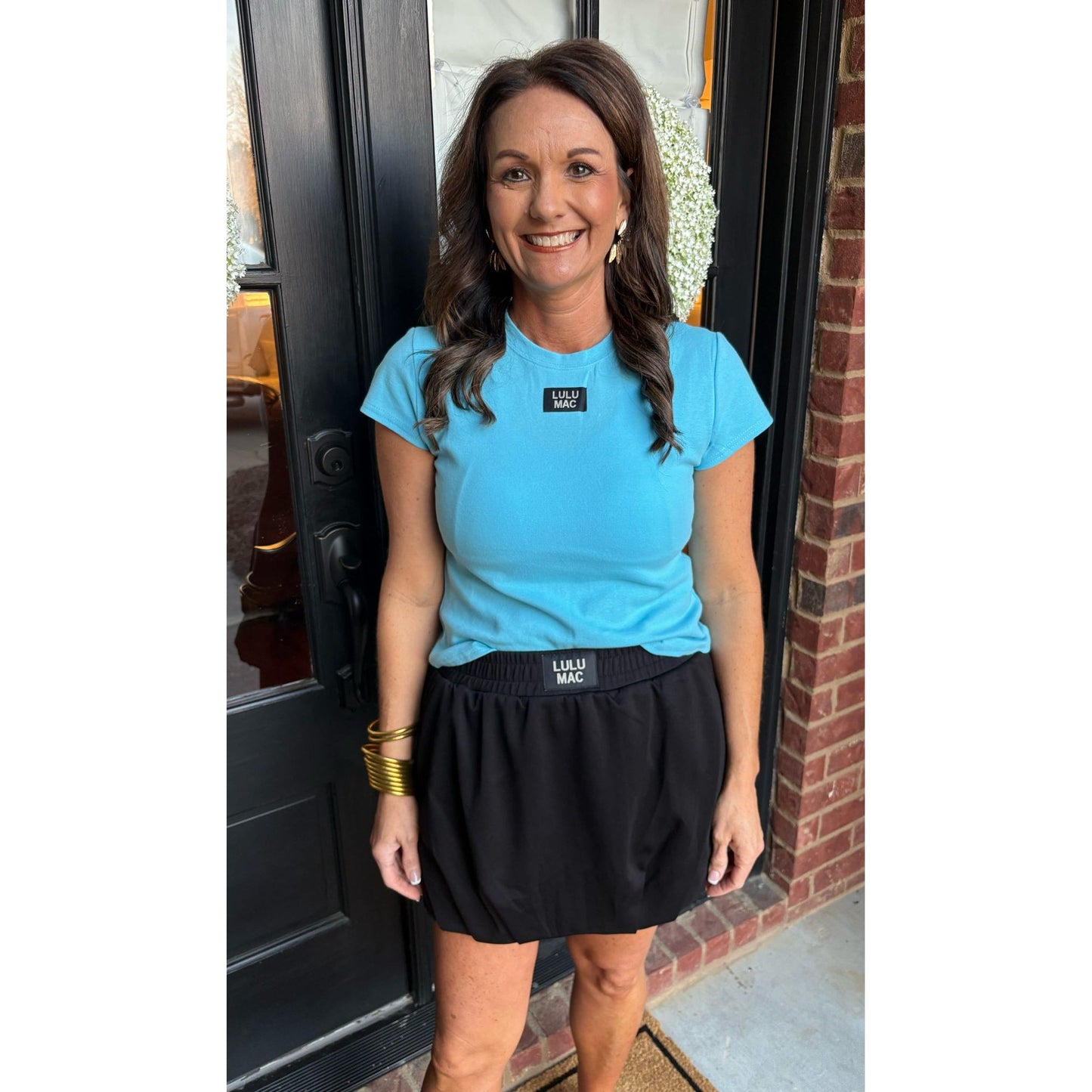 Woman wearing a blue t-shirt and black skirt standing in front of a brick wall.