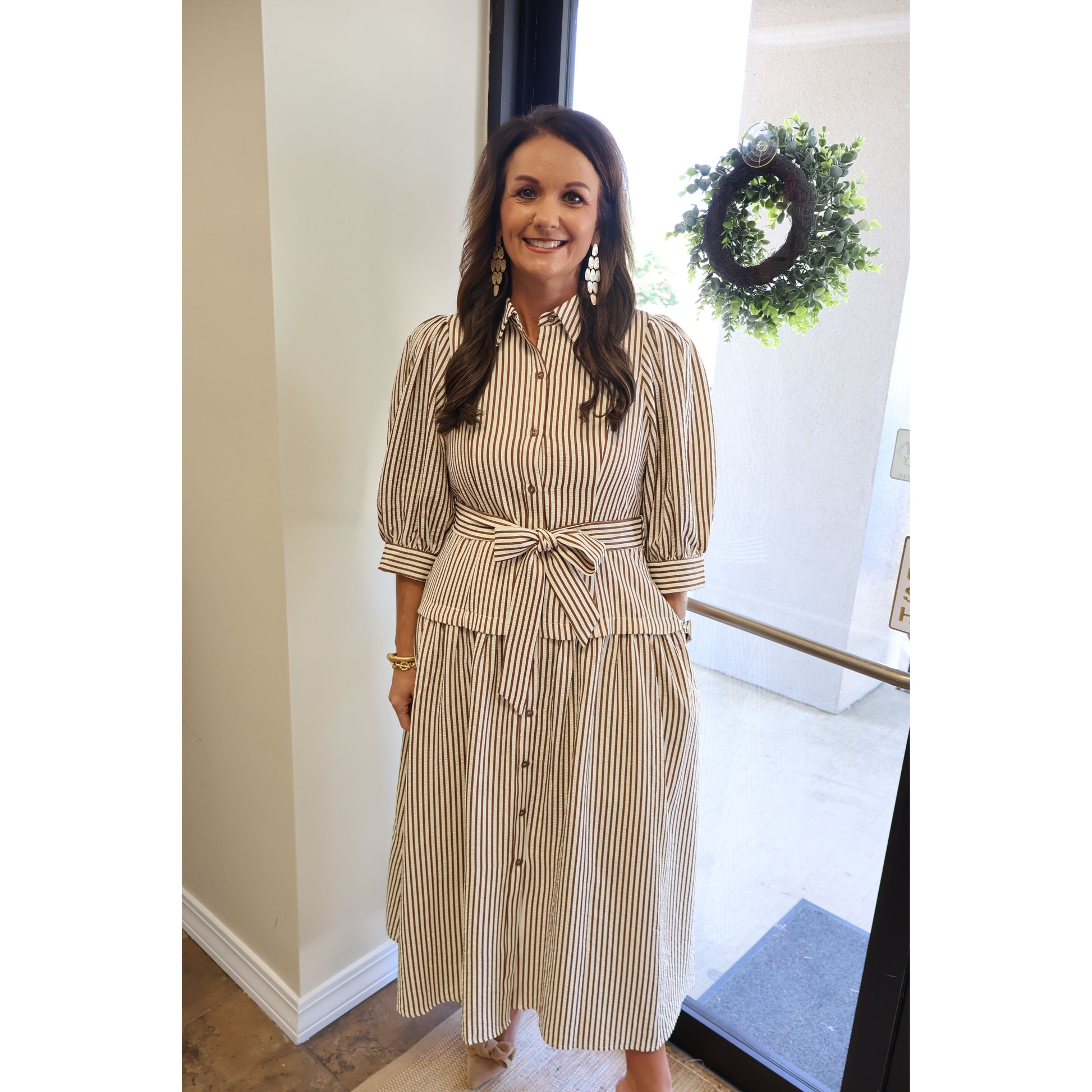A woman wearing a beige and brown vertically striped midi shirt dress with puff sleeves and a tie waist, standing in a doorway.